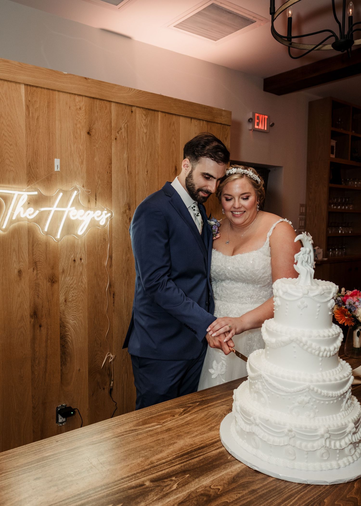 Bride and groom cut a four-tier white wedding cake beneath a custom neon sign at reception.