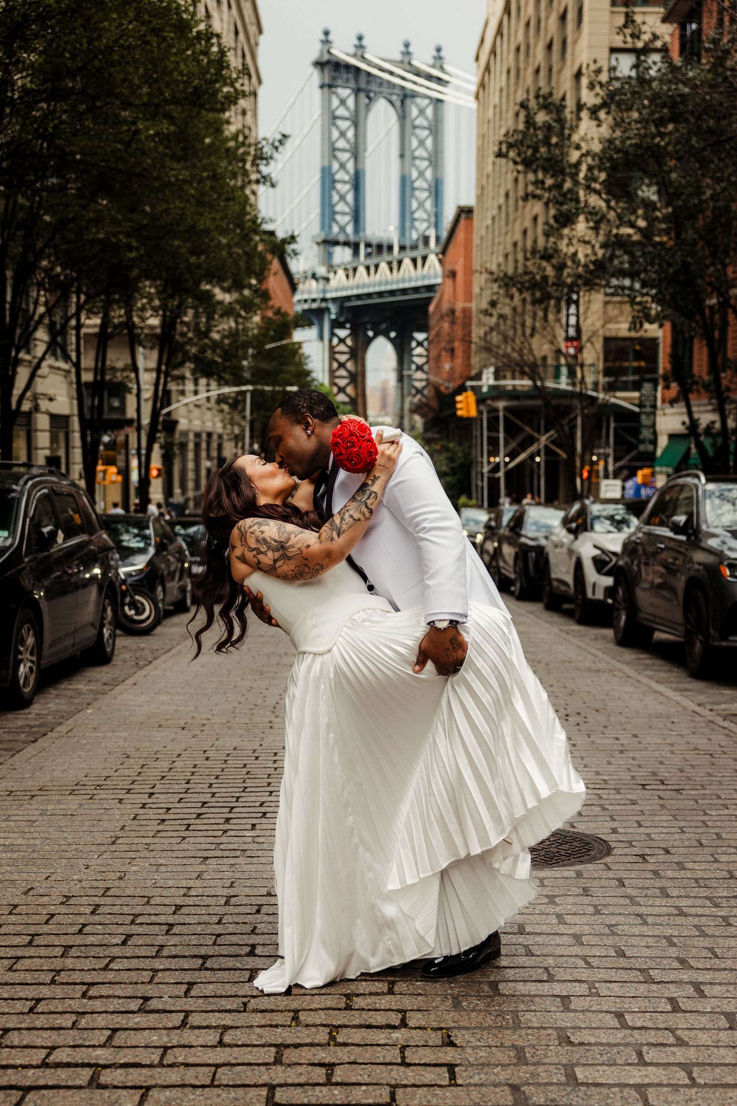 Groom dips bride for a kiss on a cobblestone street with the Manhattan Bridge in the background.