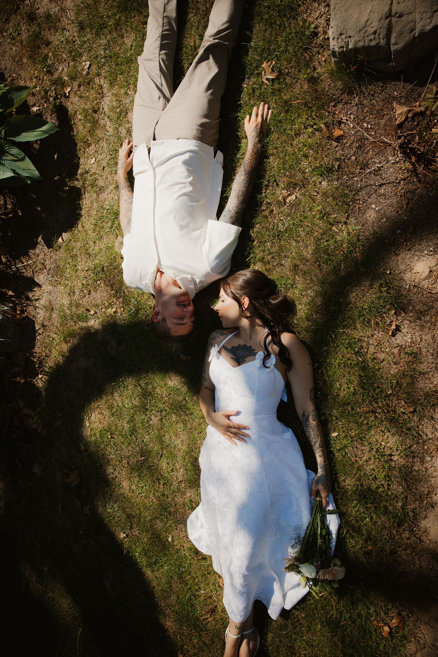 Aerial view of bride and groom lying on grass looking at each other in sunlight.