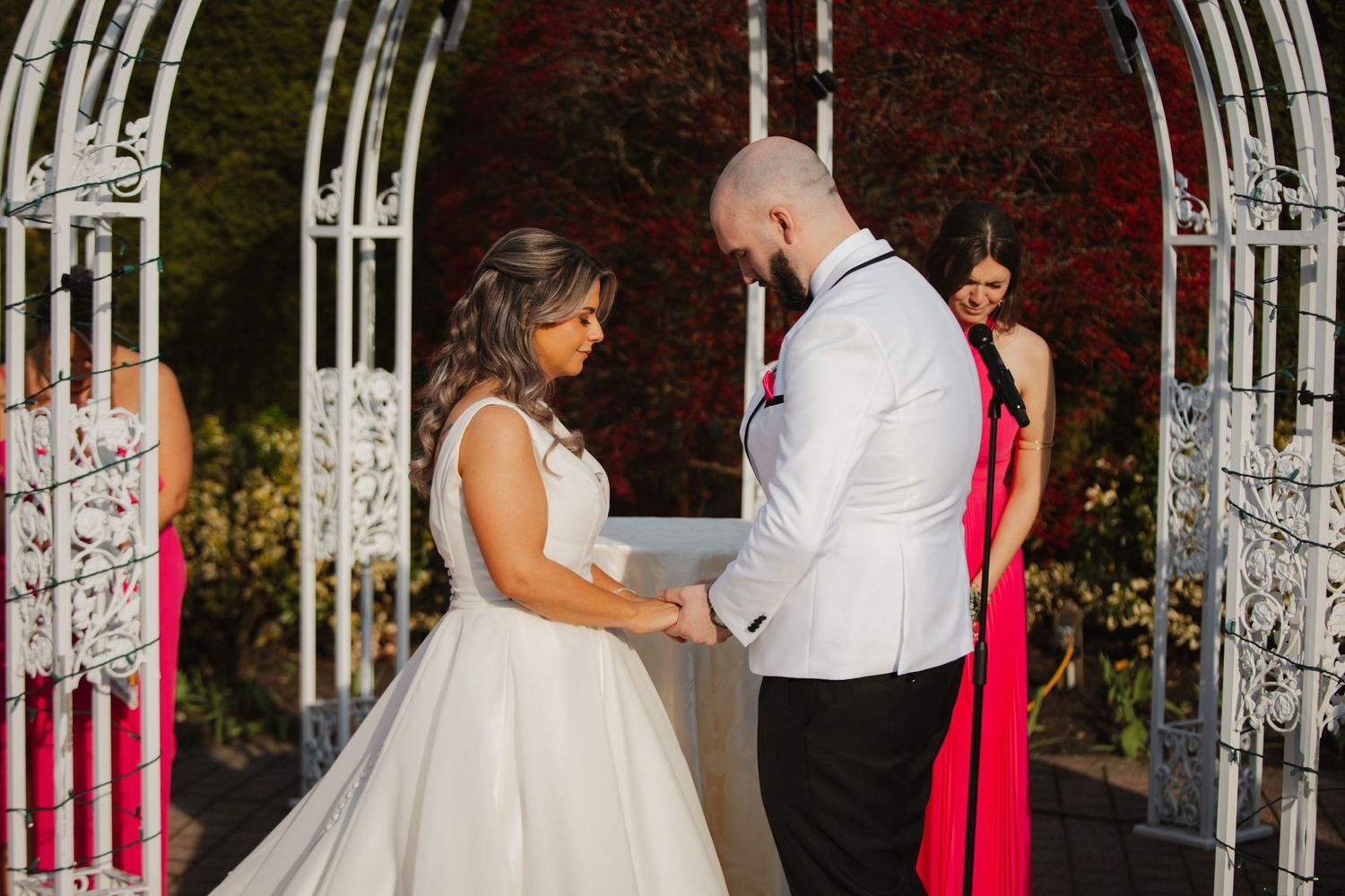 Bride and groom holding hands during outdoor wedding ceremony under white iron arch with red foliage backdrop.