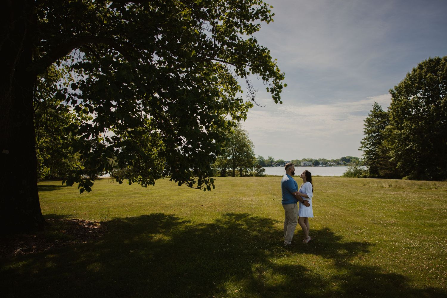 A couple walks closely together beneath a large tree with a serene lake visible in the background.