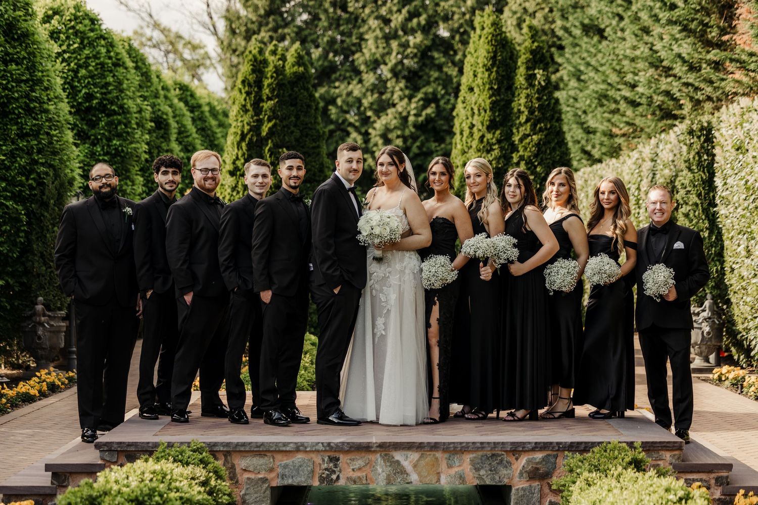 Wedding party poses in a garden with bridesmaids in black dresses and groomsmen in black suits.