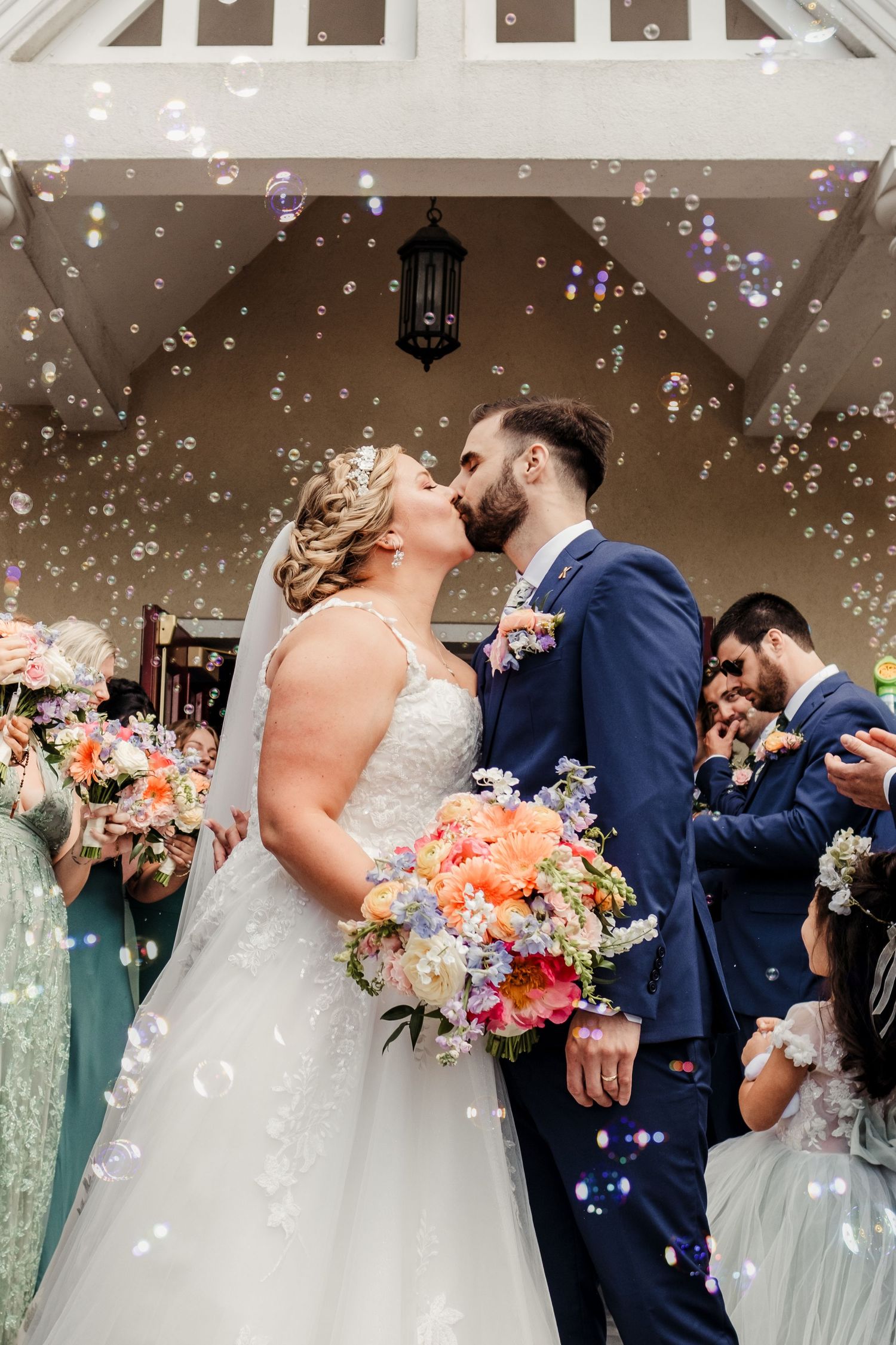 Bride and groom share first kiss surrounded by bubbles and wedding guests at ceremony exit.