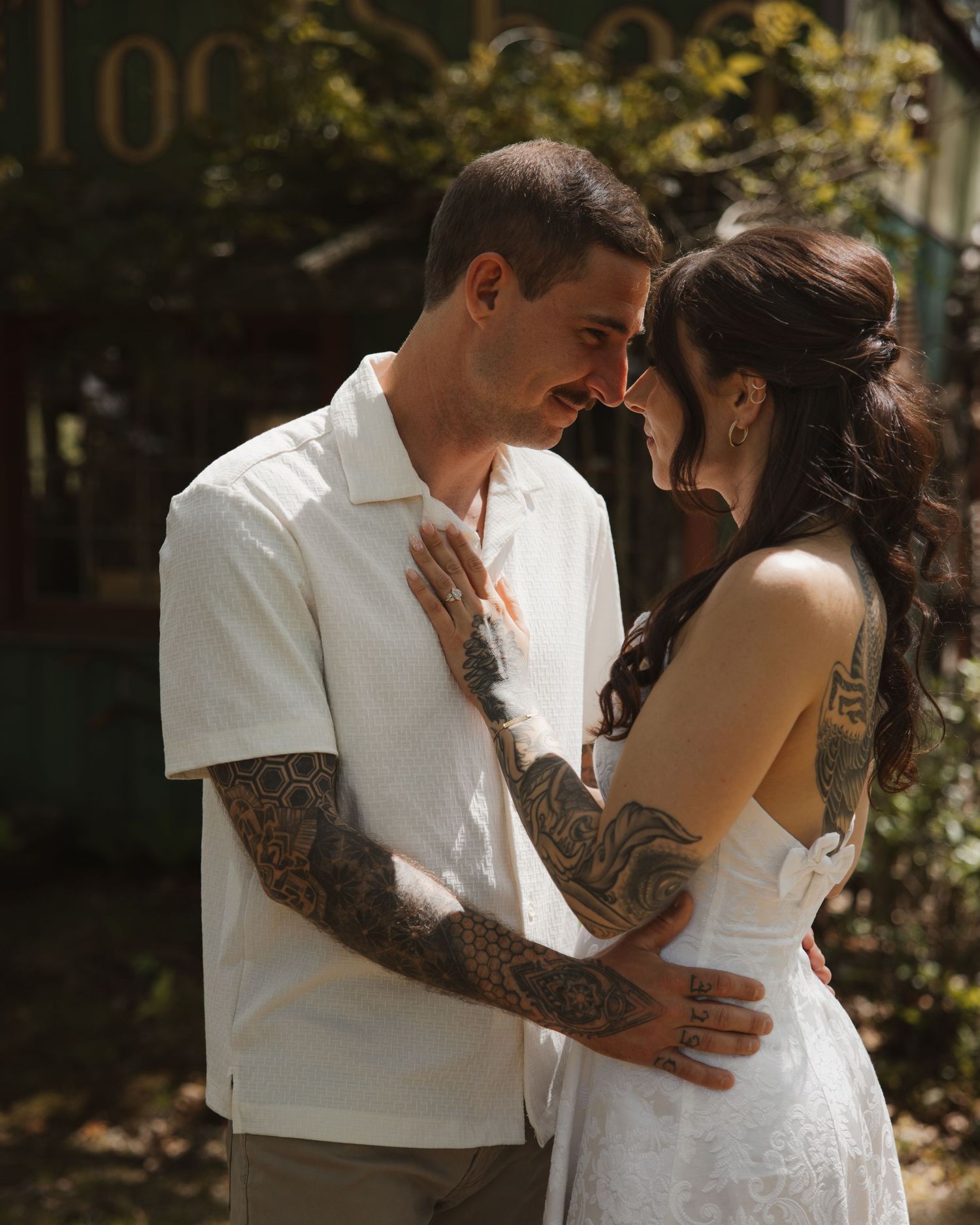 Tattooed couple shares an intimate moment outdoors, foreheads touching in a sun-dappled garden.