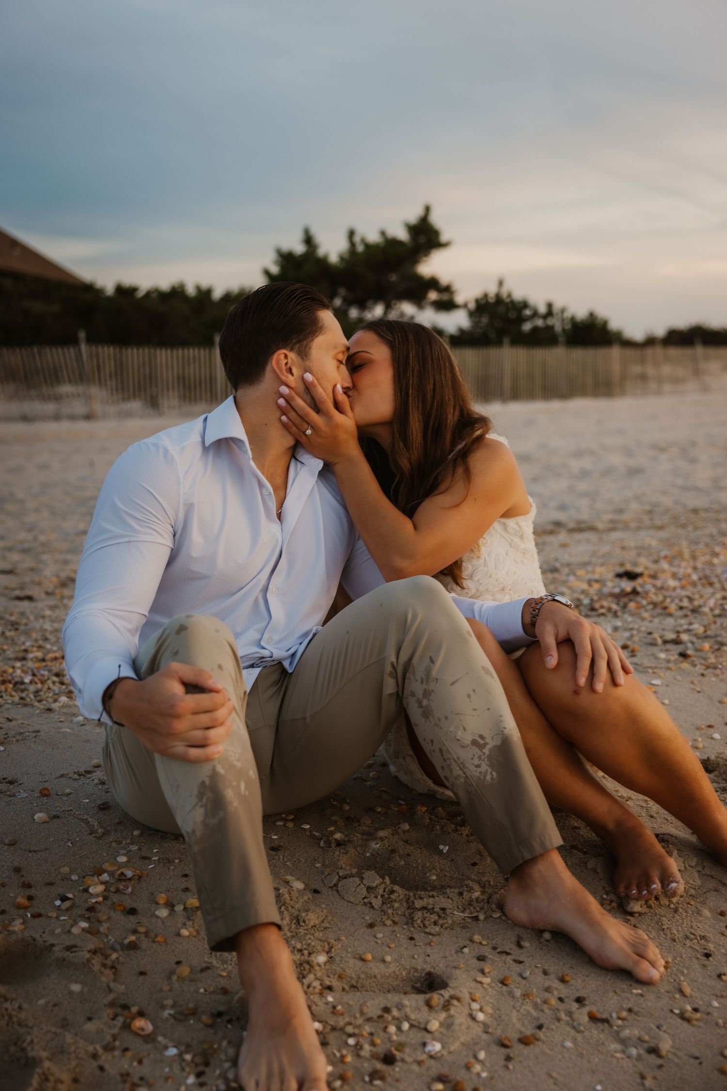 Couple sharing a romantic kiss while sitting barefoot on a sandy beach at sunset.