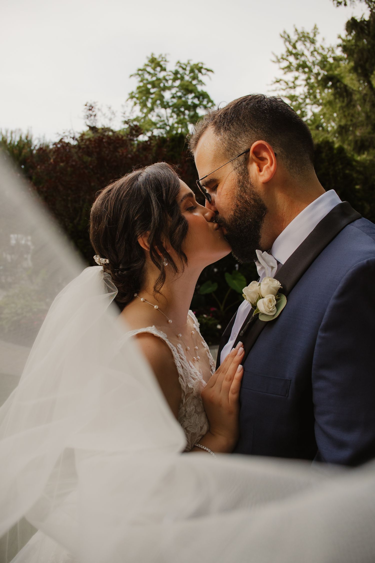 Bride and groom share a tender kiss outdoors with veil flowing in the breeze on wedding day.