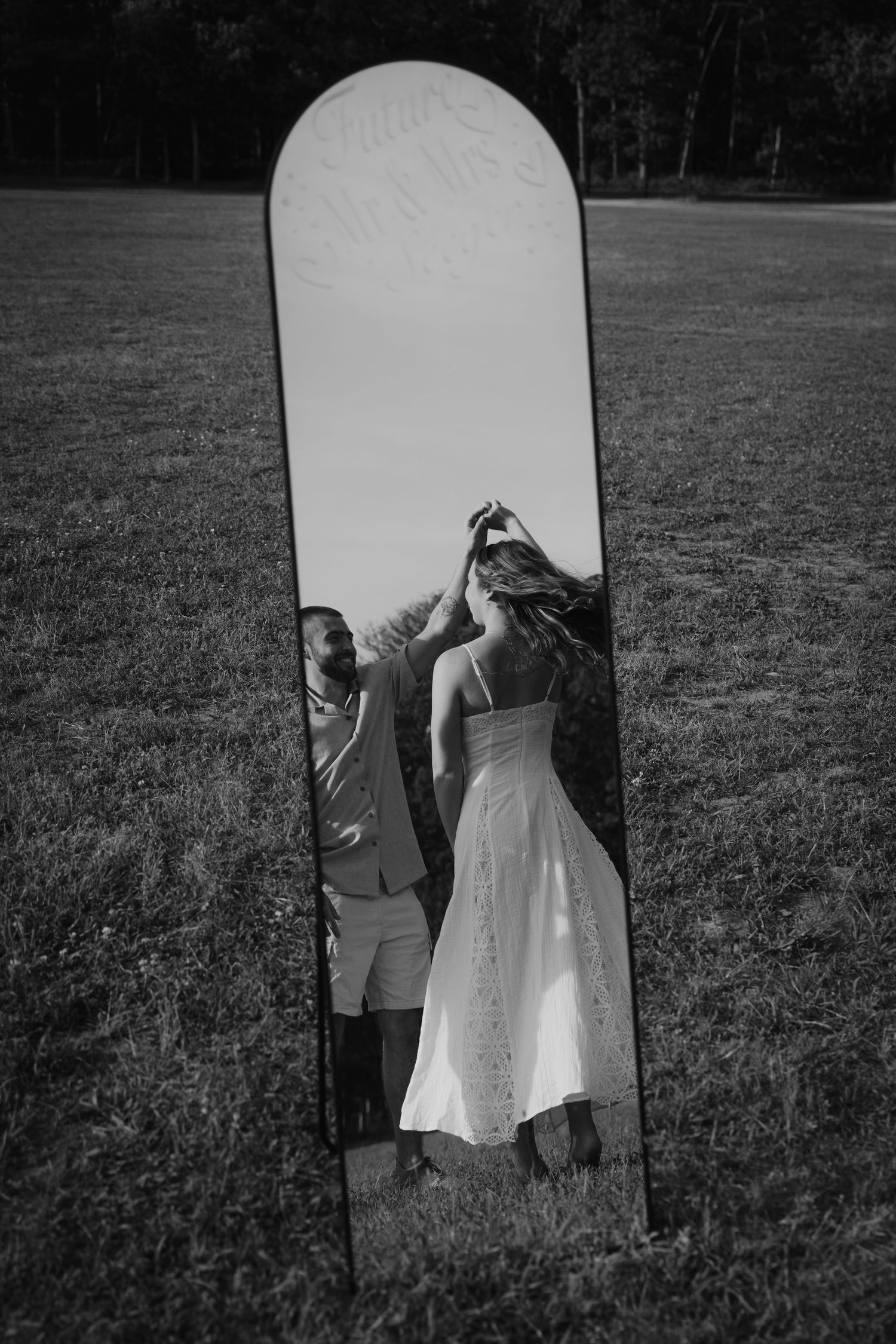Black and white reflection of a couple dancing in a tall arched mirror standing in an open grassy field.