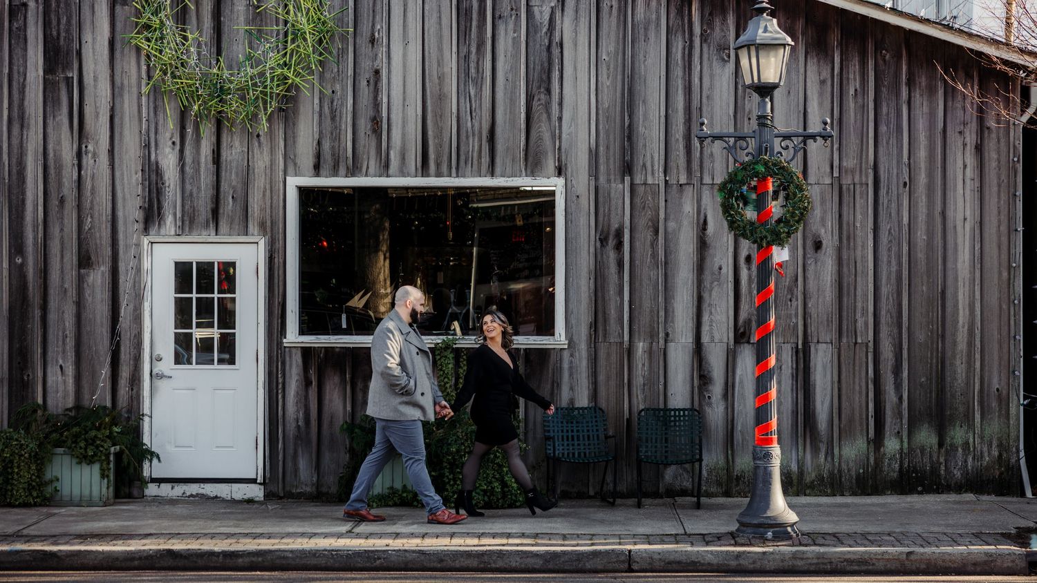 Couple walks past rustic weathered wood building with Christmas wreath decoration on lamp post.