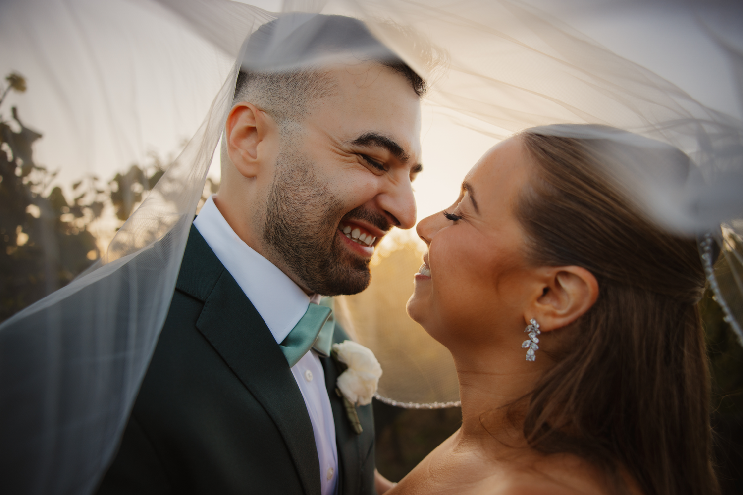 Bride and groom share a romantic moment under a veil, smiling at each other in golden sunset light.