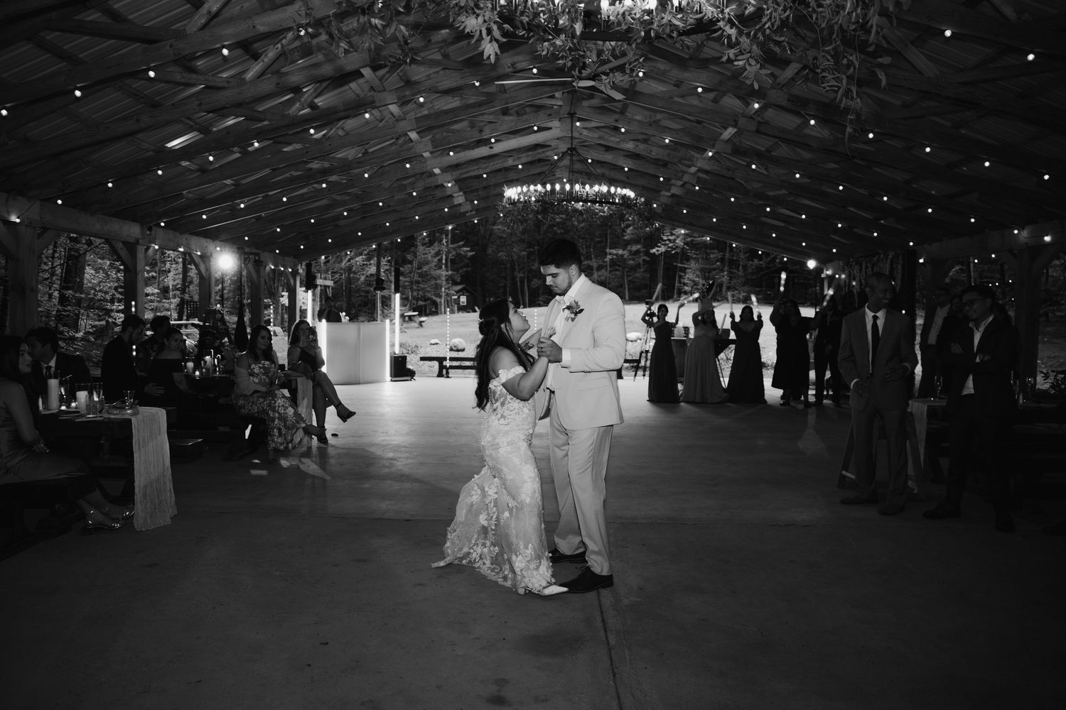 Black and white photo of bride and groom sharing first dance under twinkling lights at wedding reception.