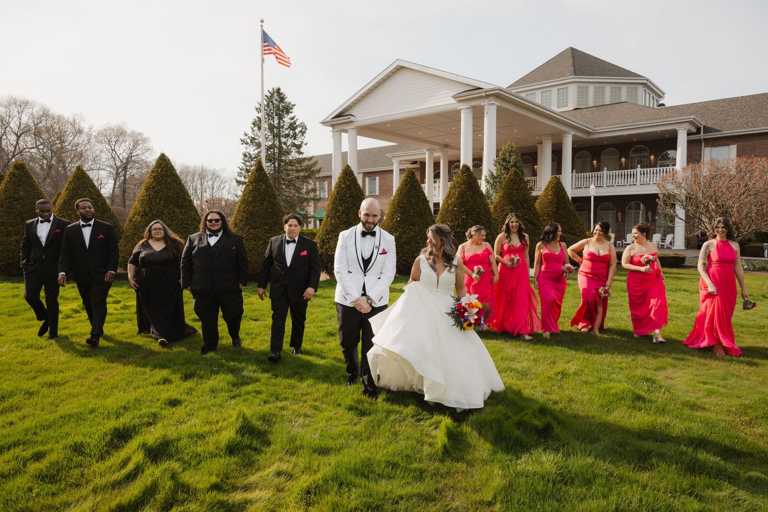 Wedding party walks across green lawn with bridesmaids in red dresses and groomsmen in black tuxedos.