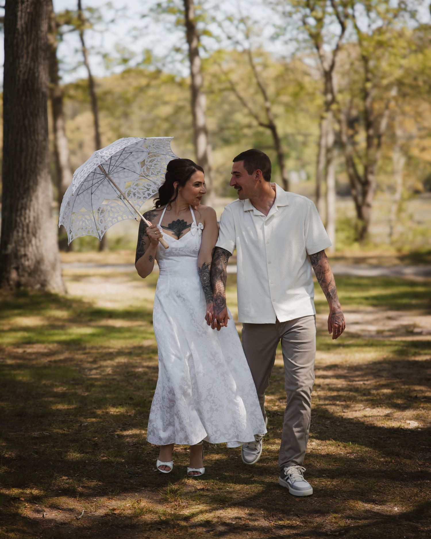 Bride and groom walk together holding a lace umbrella along a sunlit tree-lined path.