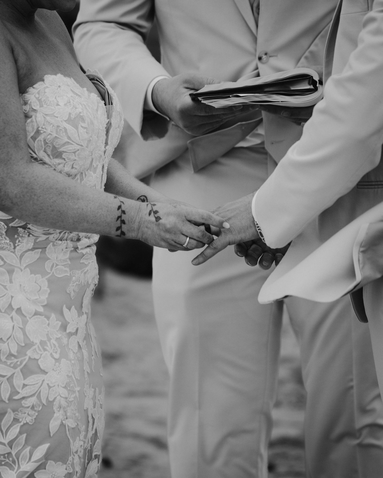Black and white close-up of bride and groom exchanging rings during beach wedding ceremony.