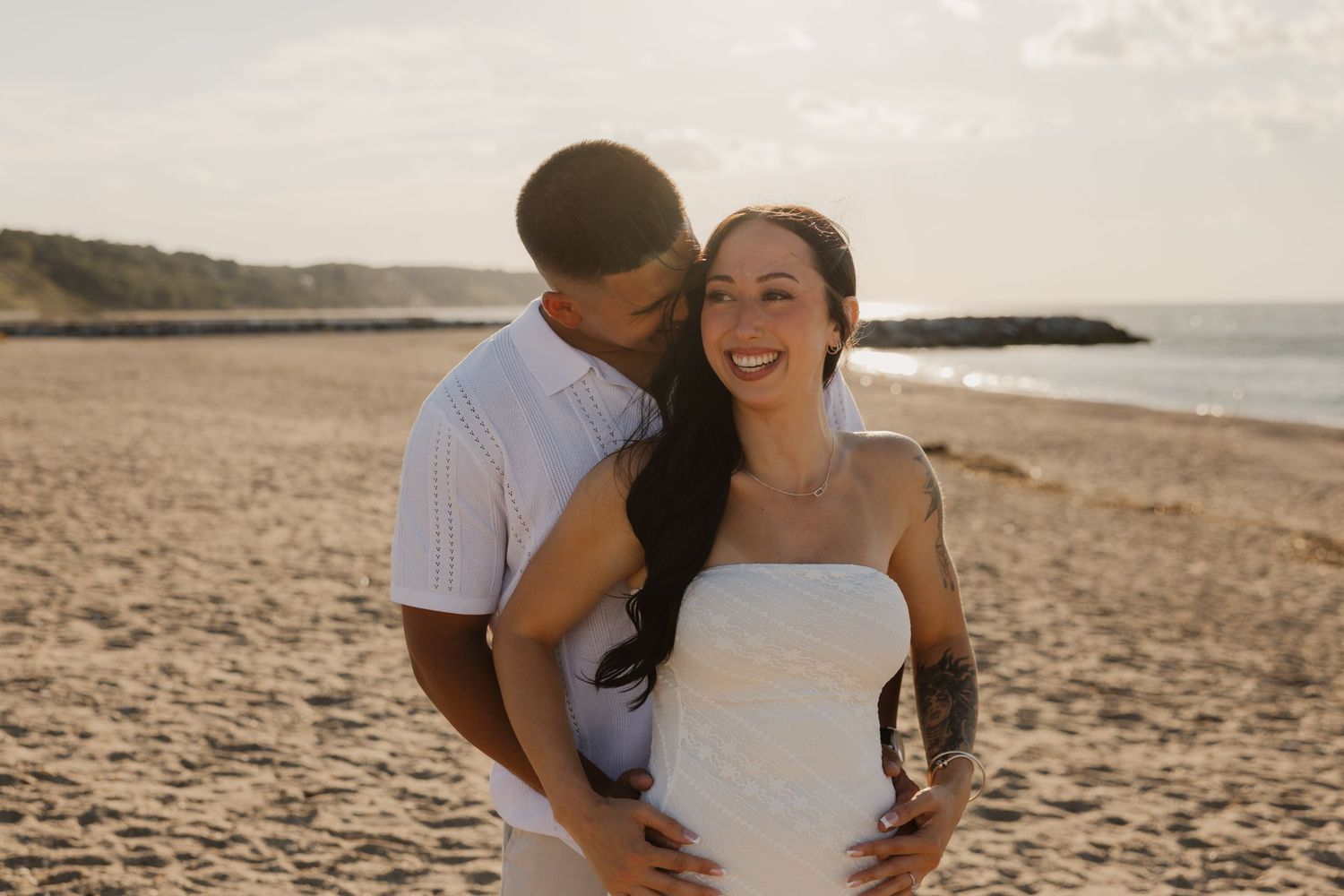 A smiling bride and groom share a tender moment on a sandy beach at sunset.