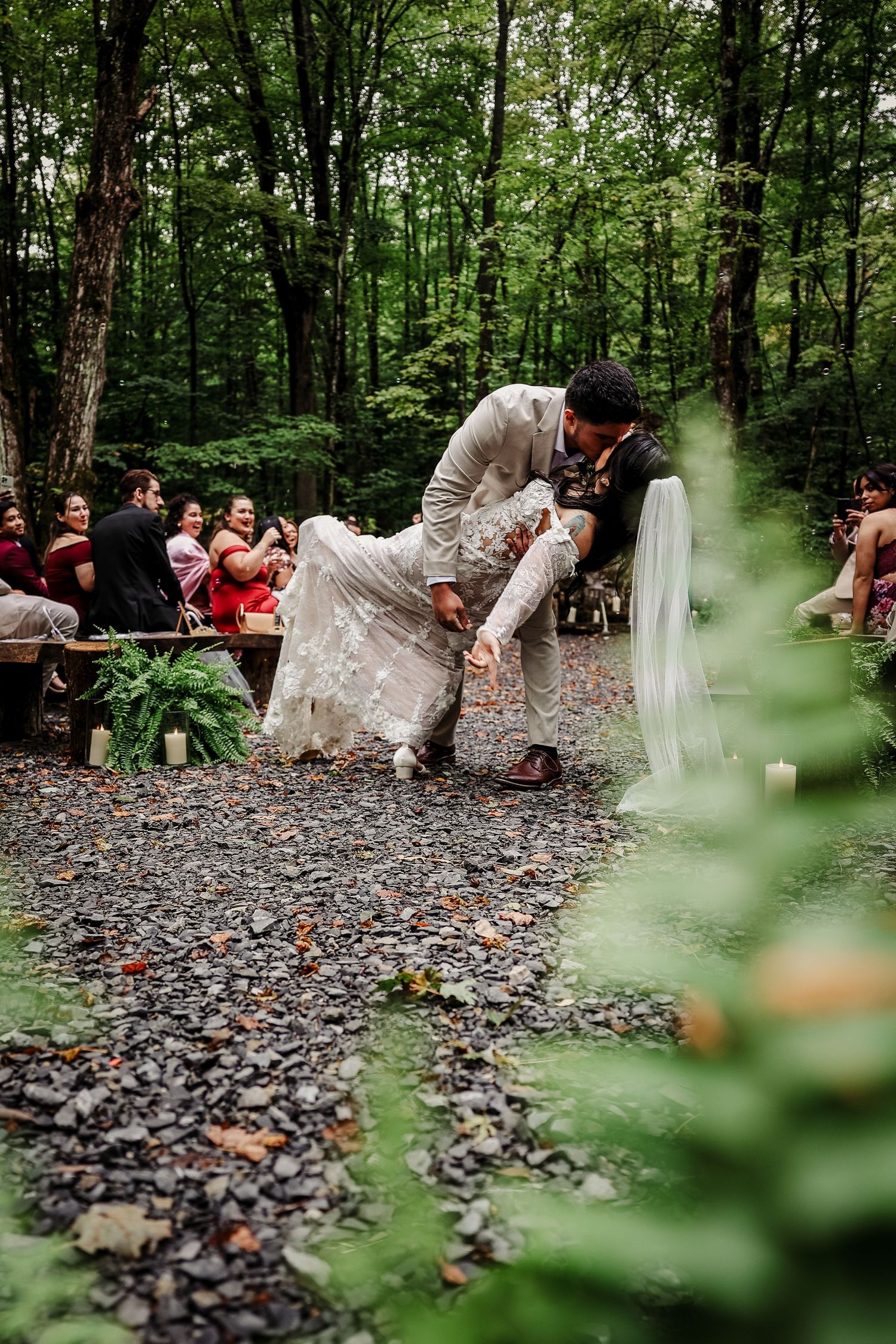Groom dips bride during outdoor woodland wedding ceremony on gravel path with guests seated among trees.