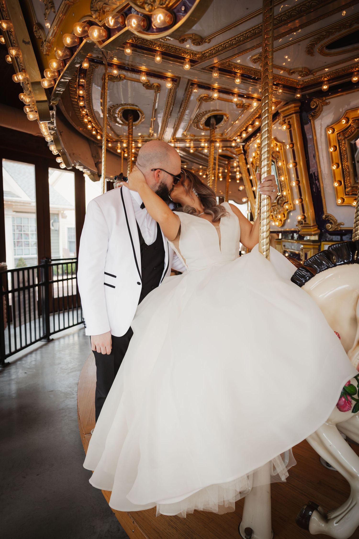 Bride and groom share a romantic kiss on a vintage carousel with golden lights and ornate details.