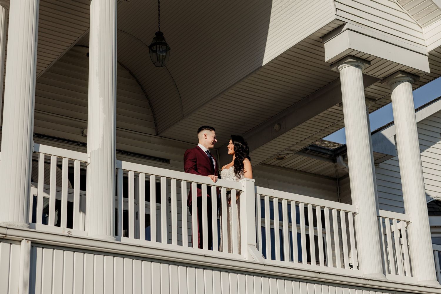 Couple stands together on a white colonial porch balcony during golden hour at their wedding venue.