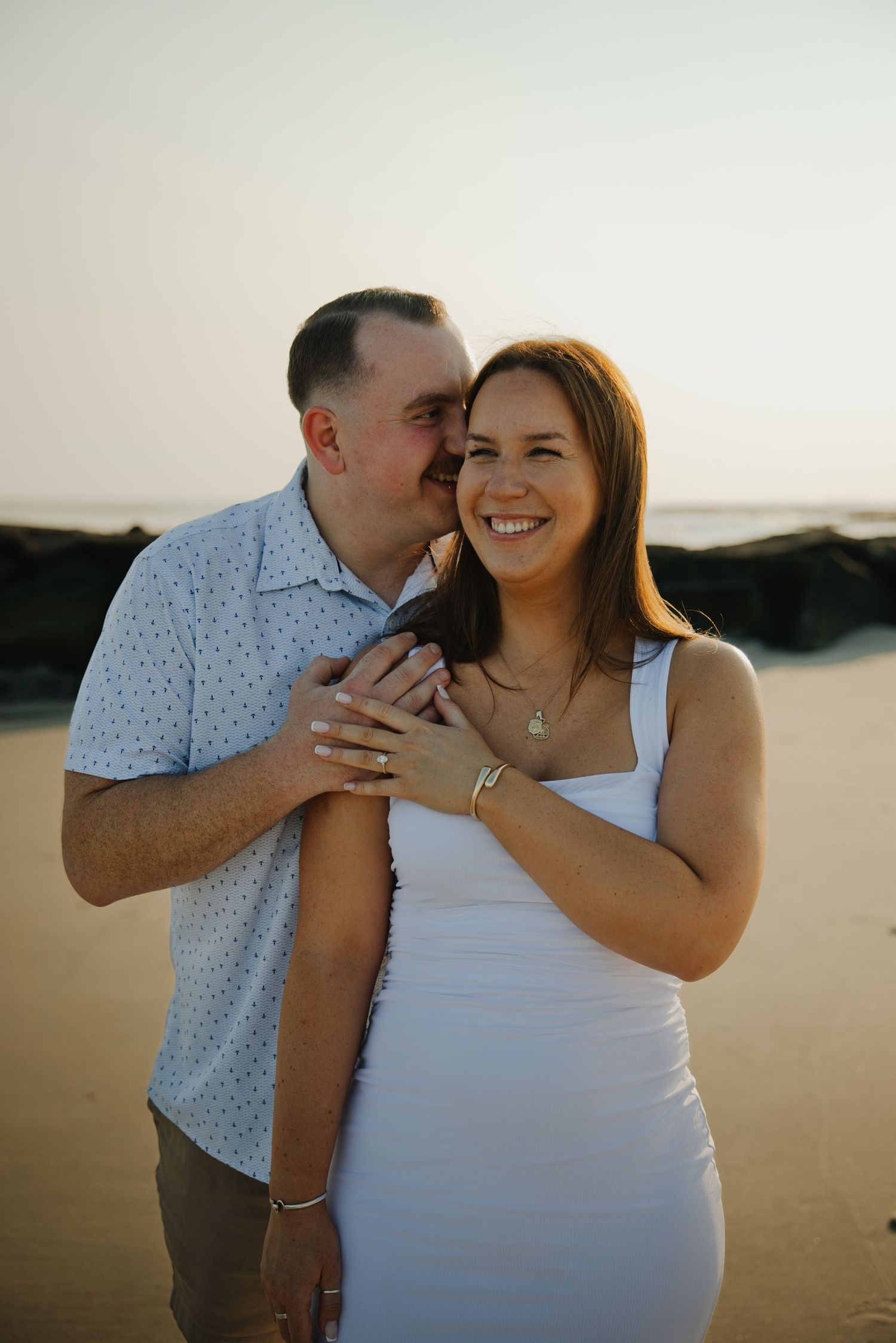 Man kissing woman on cheek from behind as she smiles on beach at golden hour sunset.