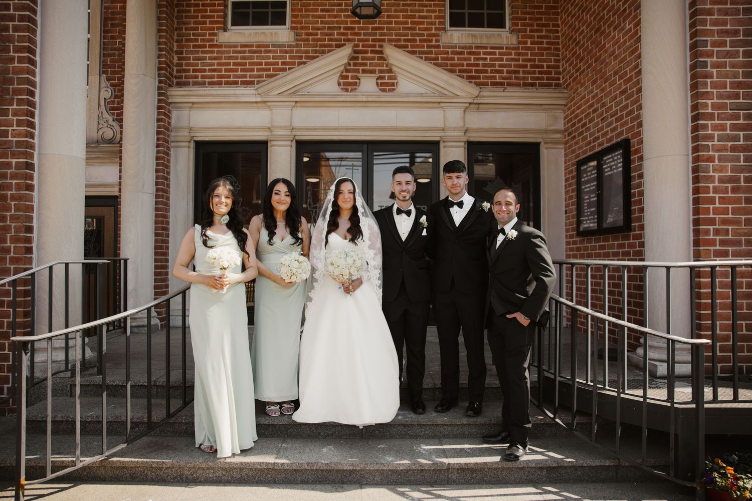 Wedding party poses on church steps with bride in white gown and groom in black tuxedo.