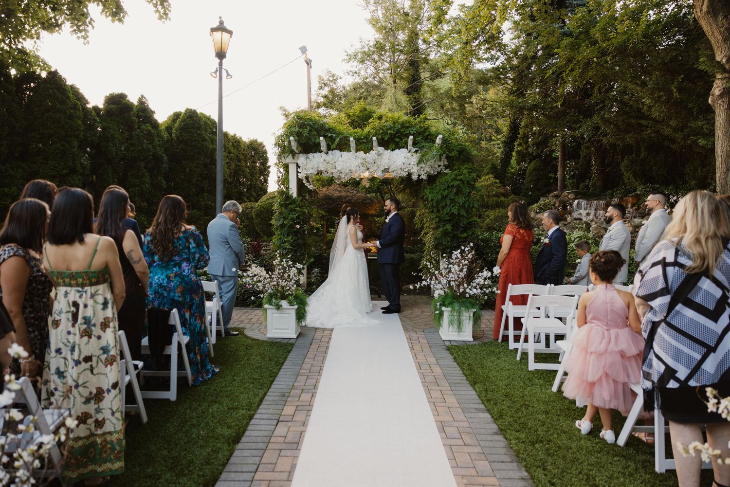Outdoor wedding ceremony with guests seated along white aisle leading to floral arch at dusk.