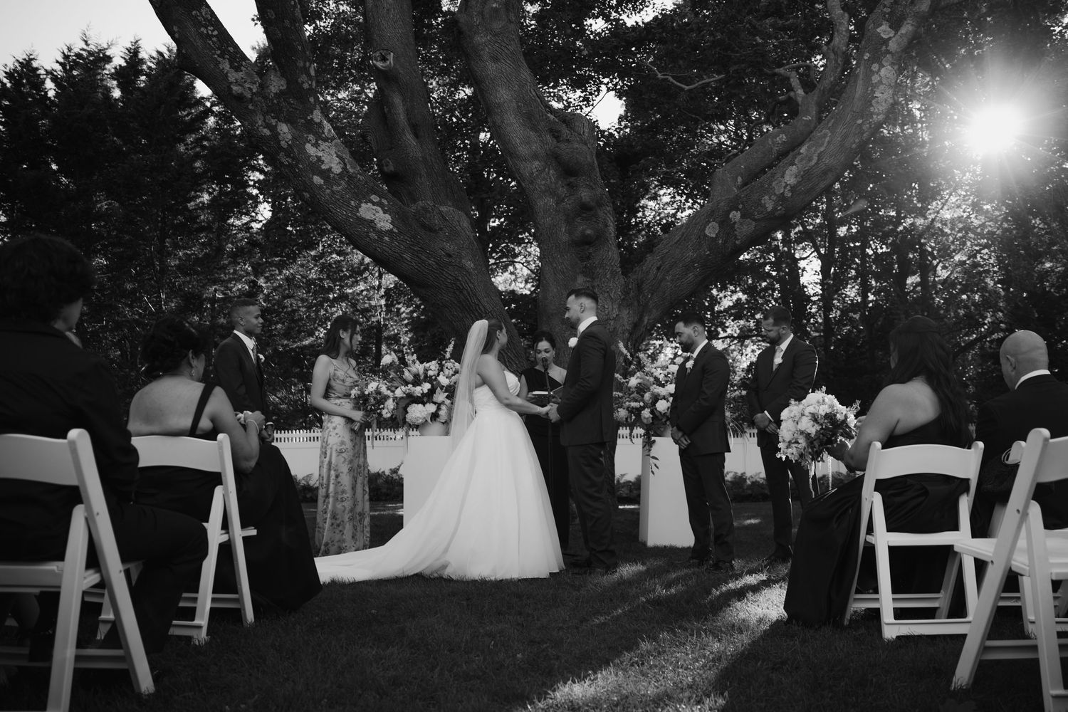 Black and white outdoor wedding ceremony under a large tree with bride, groom, and guests seated in white chairs.