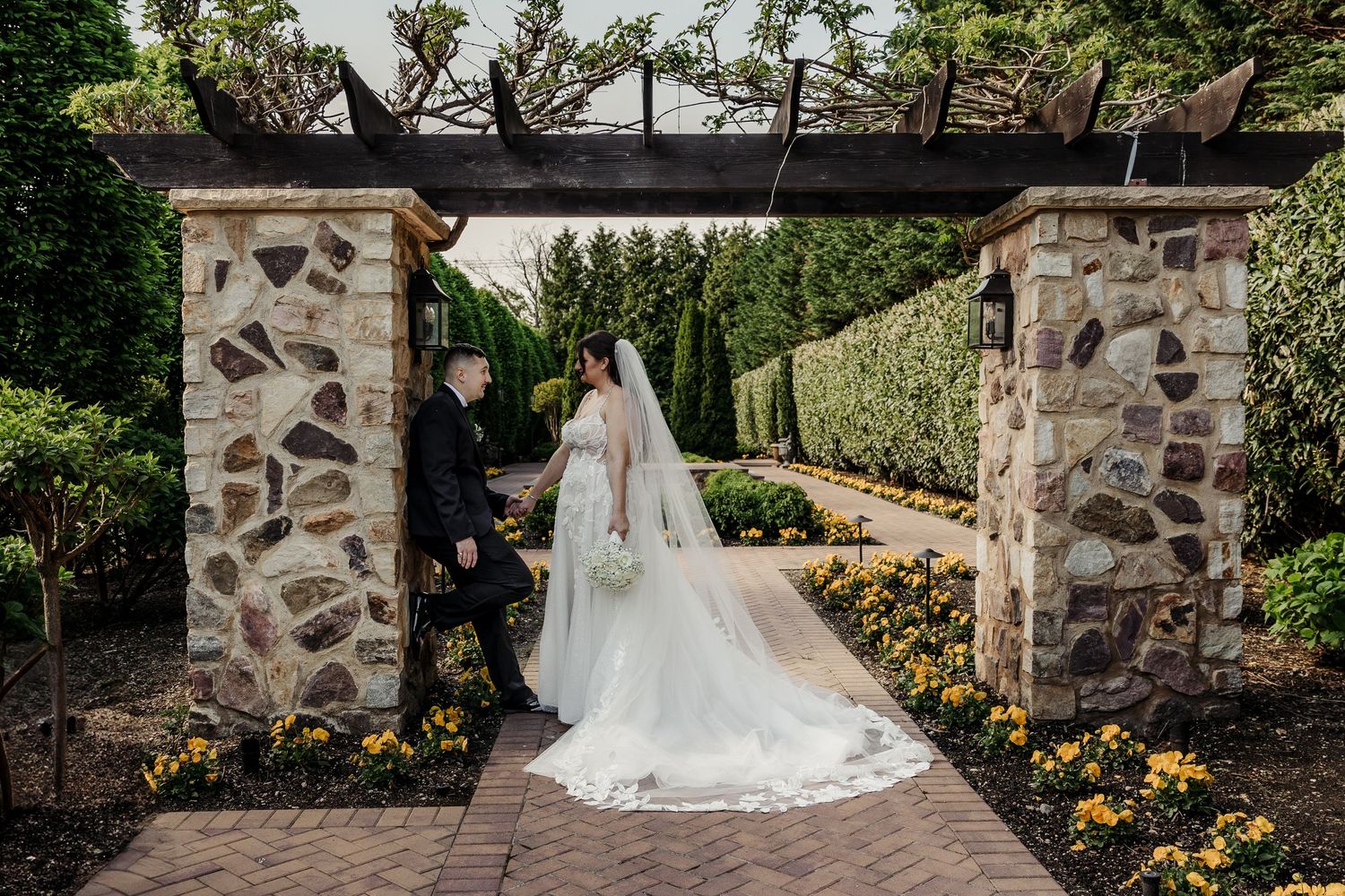 Bride and groom share a romantic moment under a stone pergola in a lush garden wedding venue.