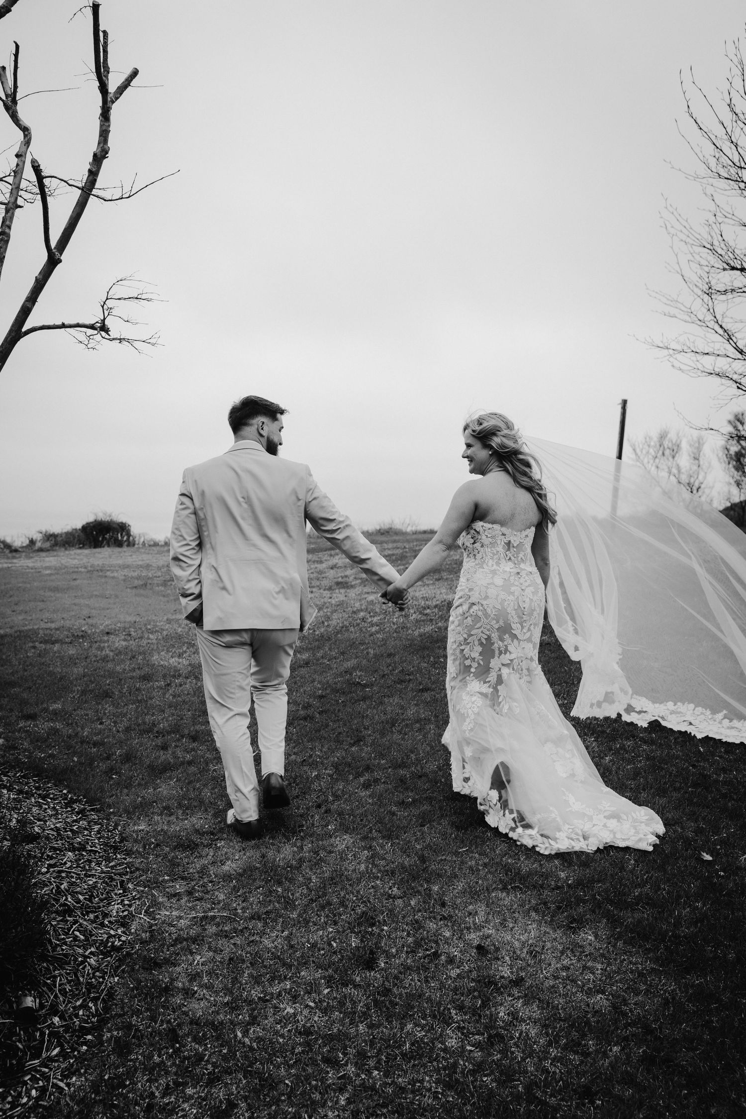 Black and white photo of bride and groom holding hands walking through field with veil flowing.