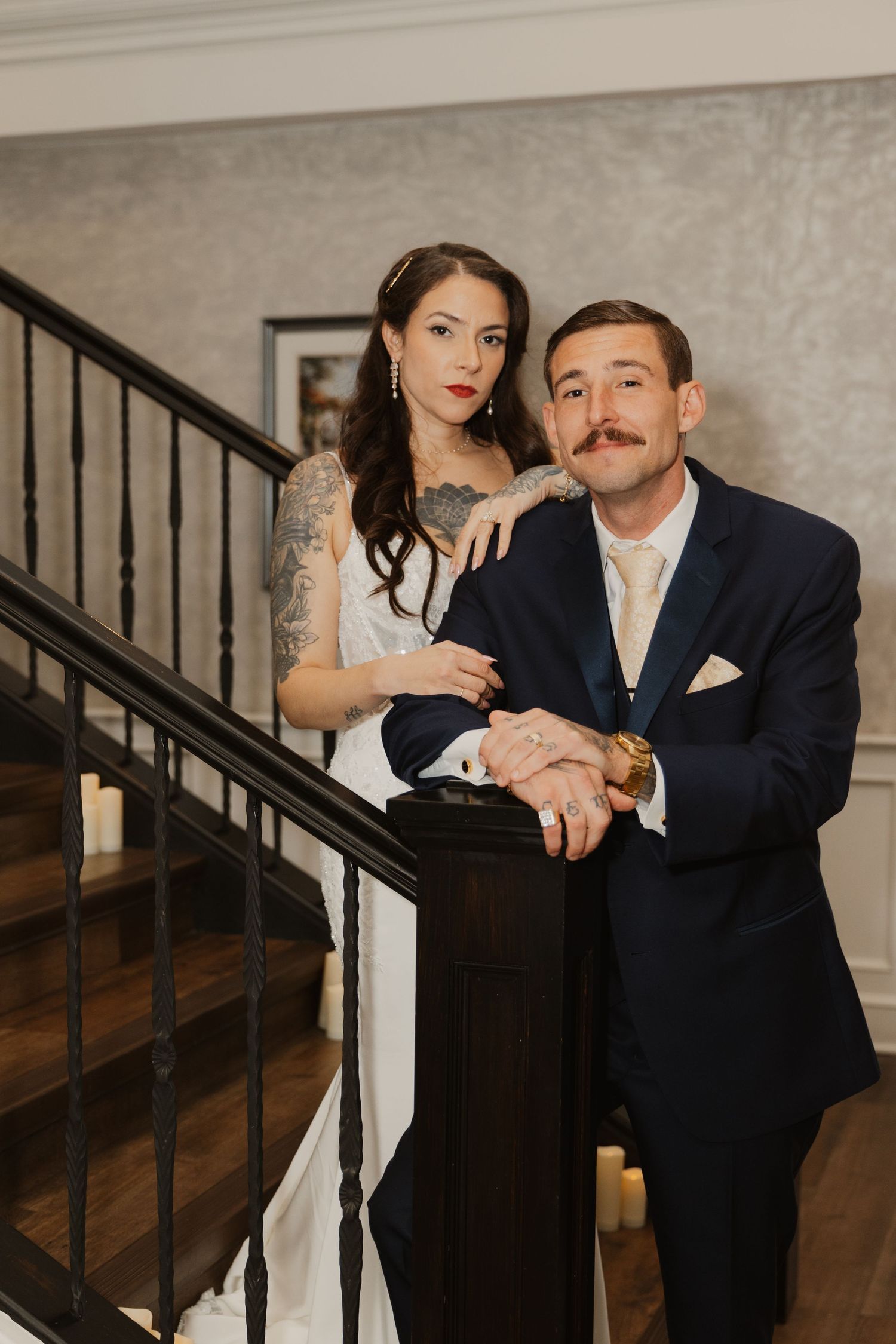 Bride with tattooed arms and groom in navy suit with mustache pose together on a dark wooden staircase with candles at an elegant wedding.