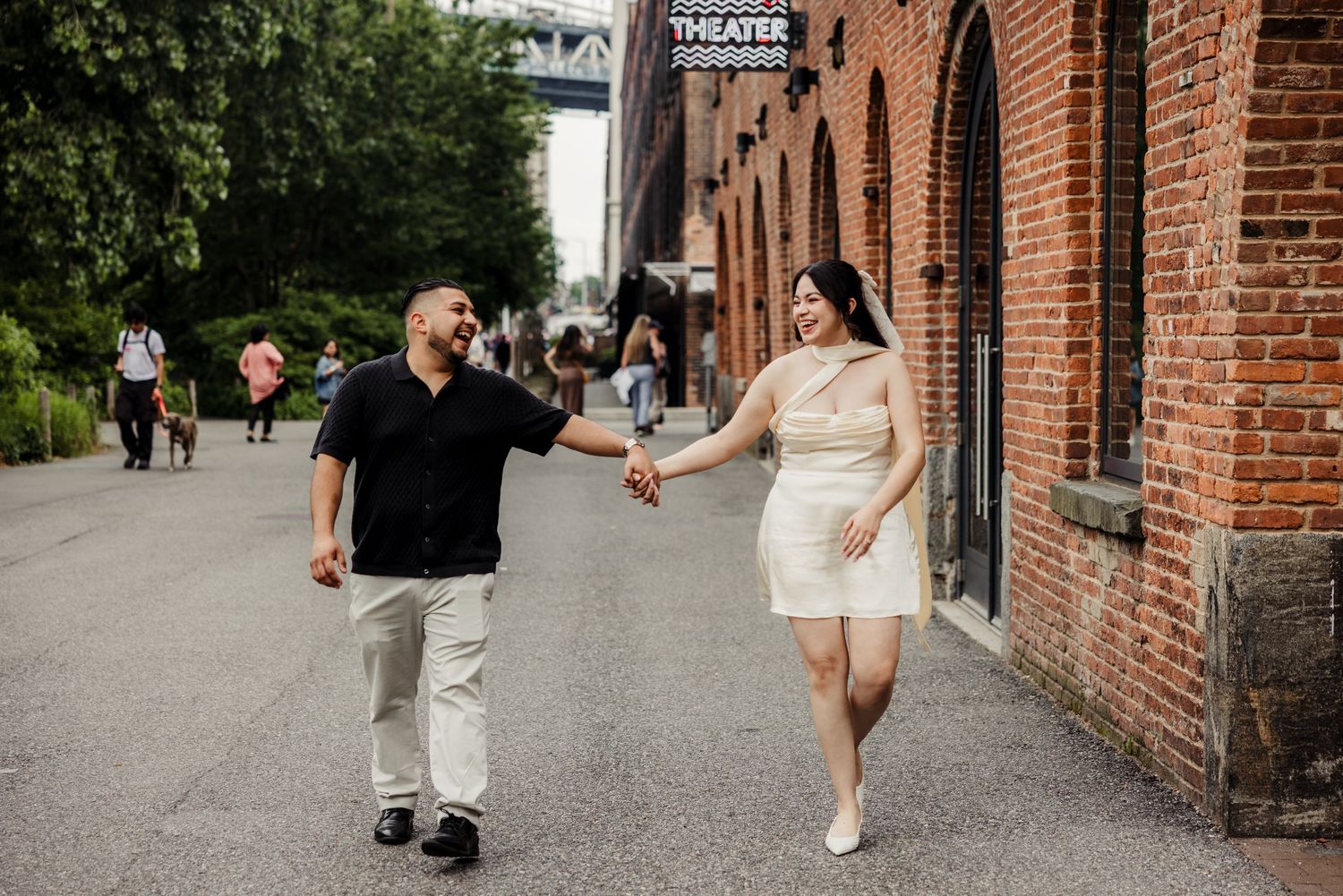 Couple walks hand-in-hand along brick building path, woman in white dress and man in black shirt smiling.