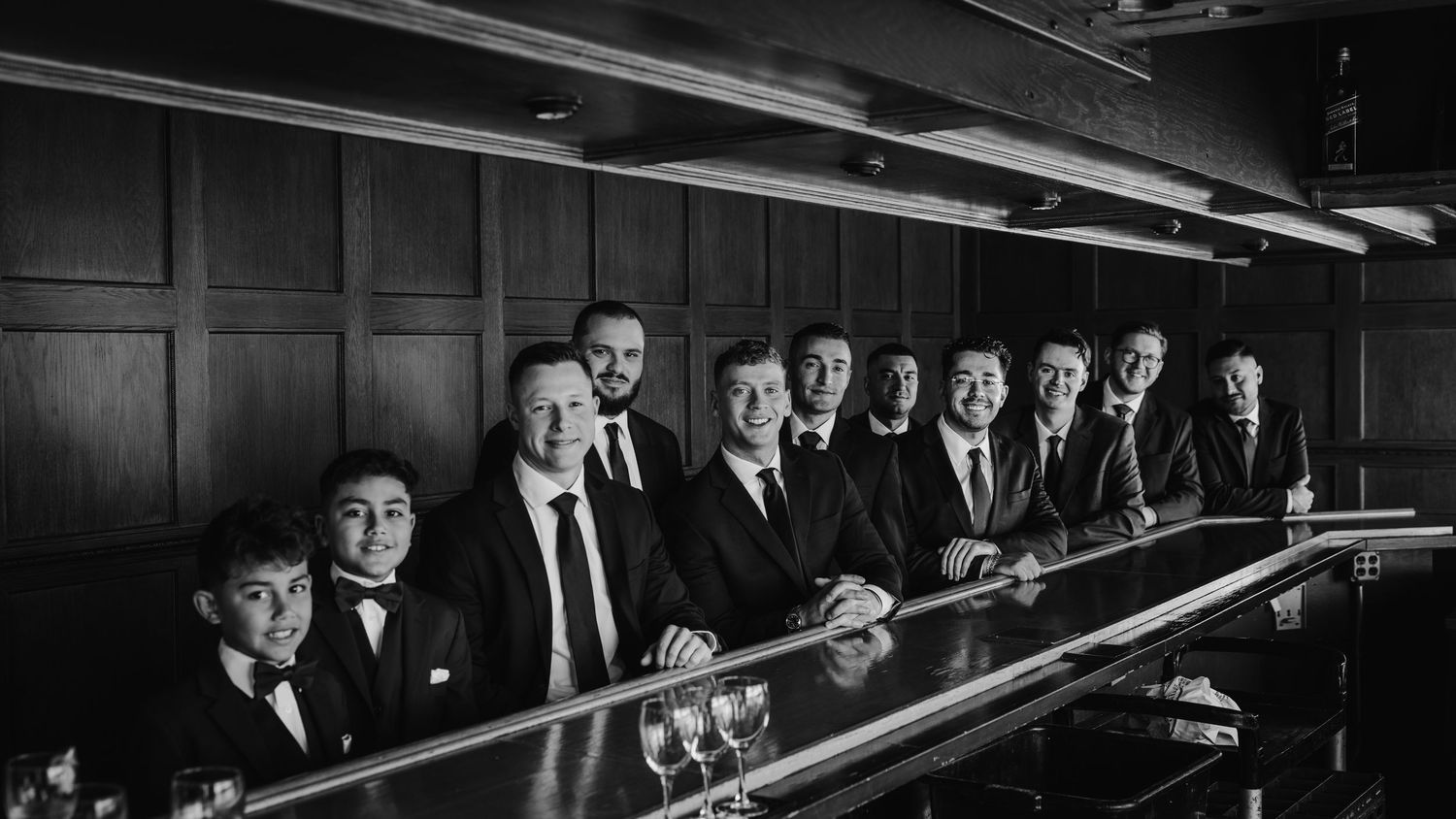 Groomsmen in black suits pose in a line at a bar in a classic black and white wedding photo.
