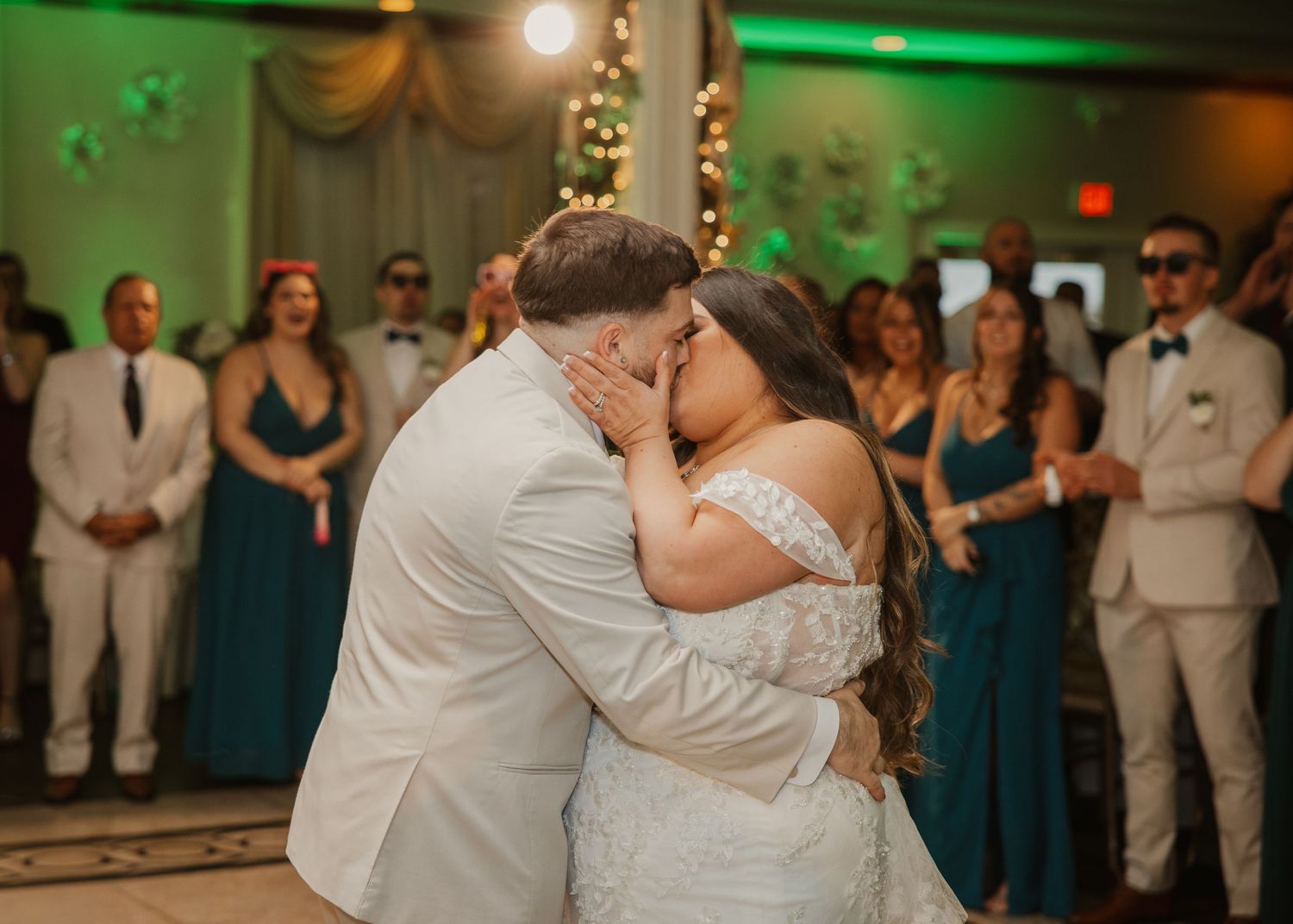 Bride and groom share passionate first kiss on dance floor surrounded by wedding guests.