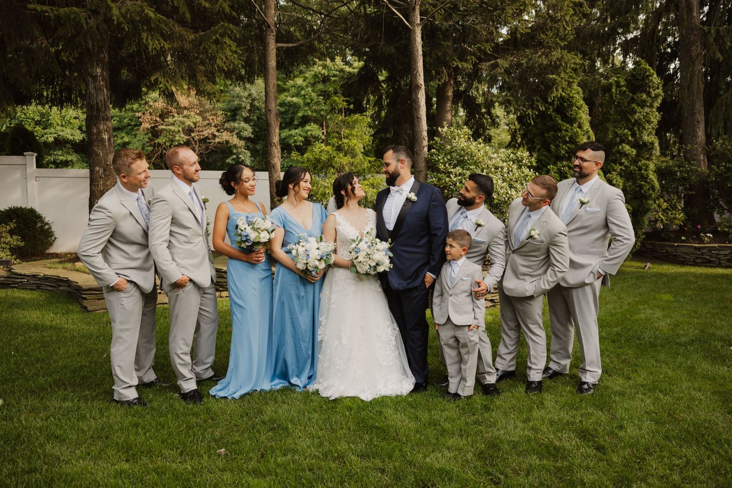 Wedding party poses on green lawn, bride in white gown with bridesmaids in blue dresses and groomsmen in gray suits.