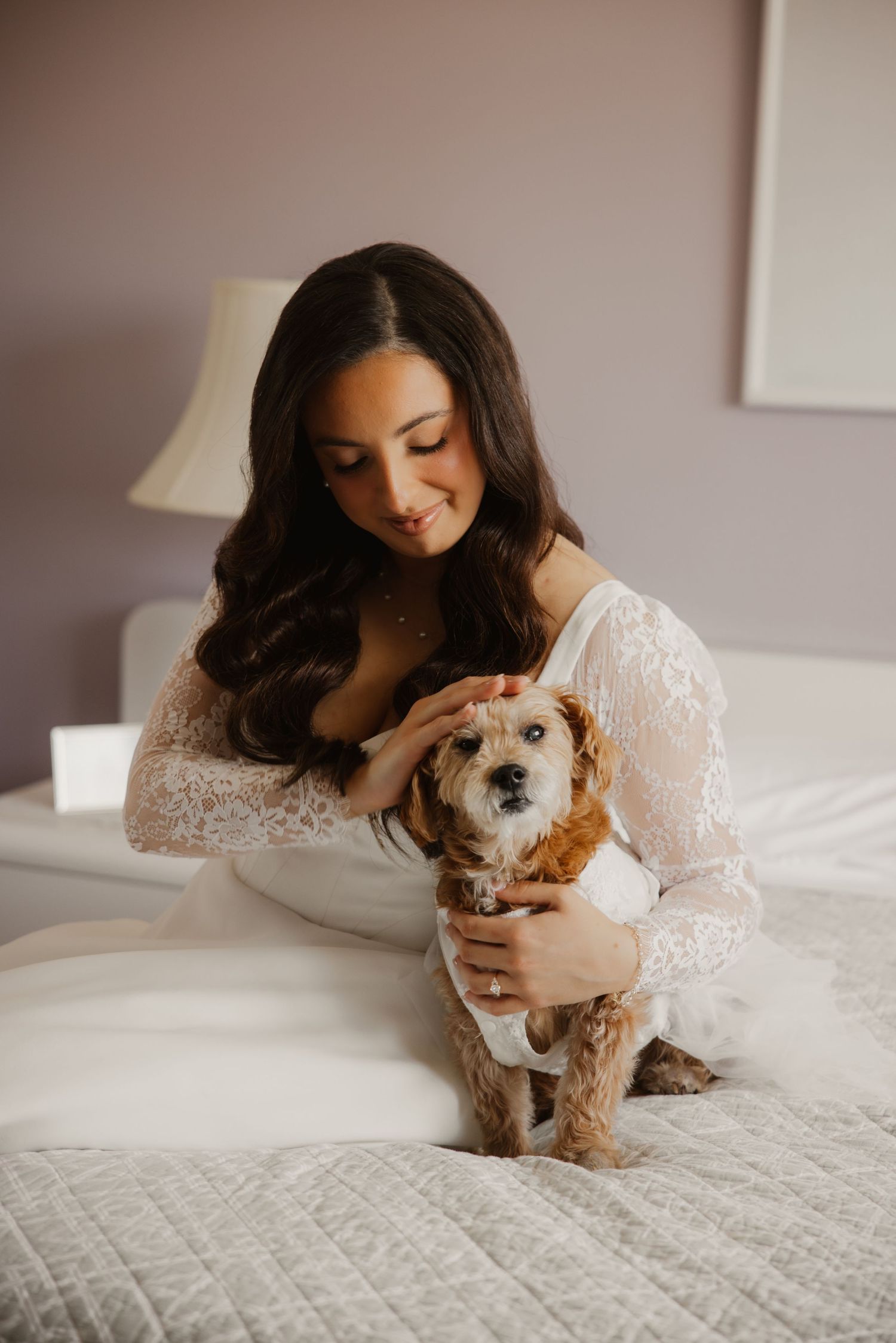 Beautiful dark-haired bride in lace robe tenderly holds her small fluffy dog on a white bed.