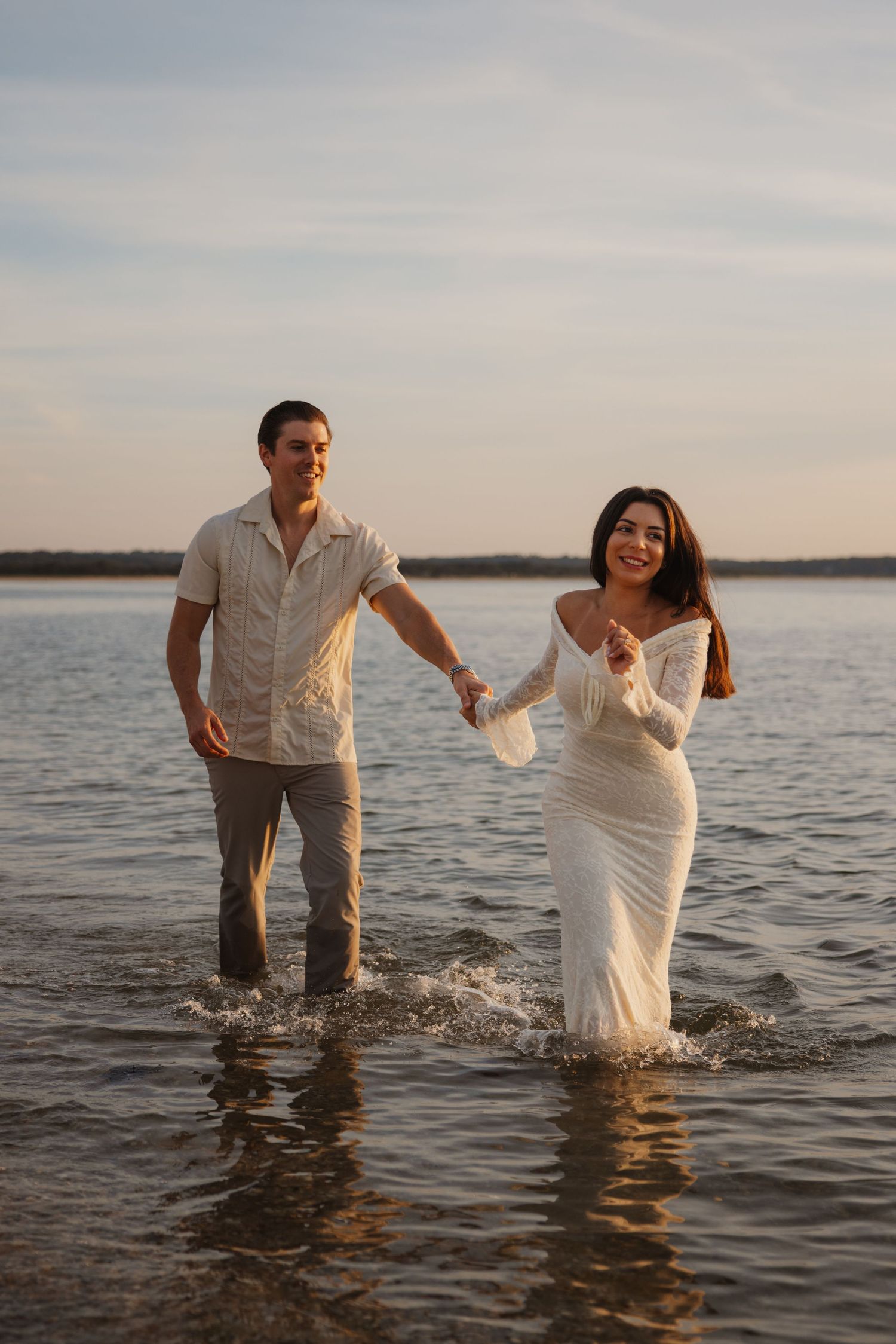 Couple holding hands wading through shallow water at sunset, woman in white dress, man in light shirt and gray pants.