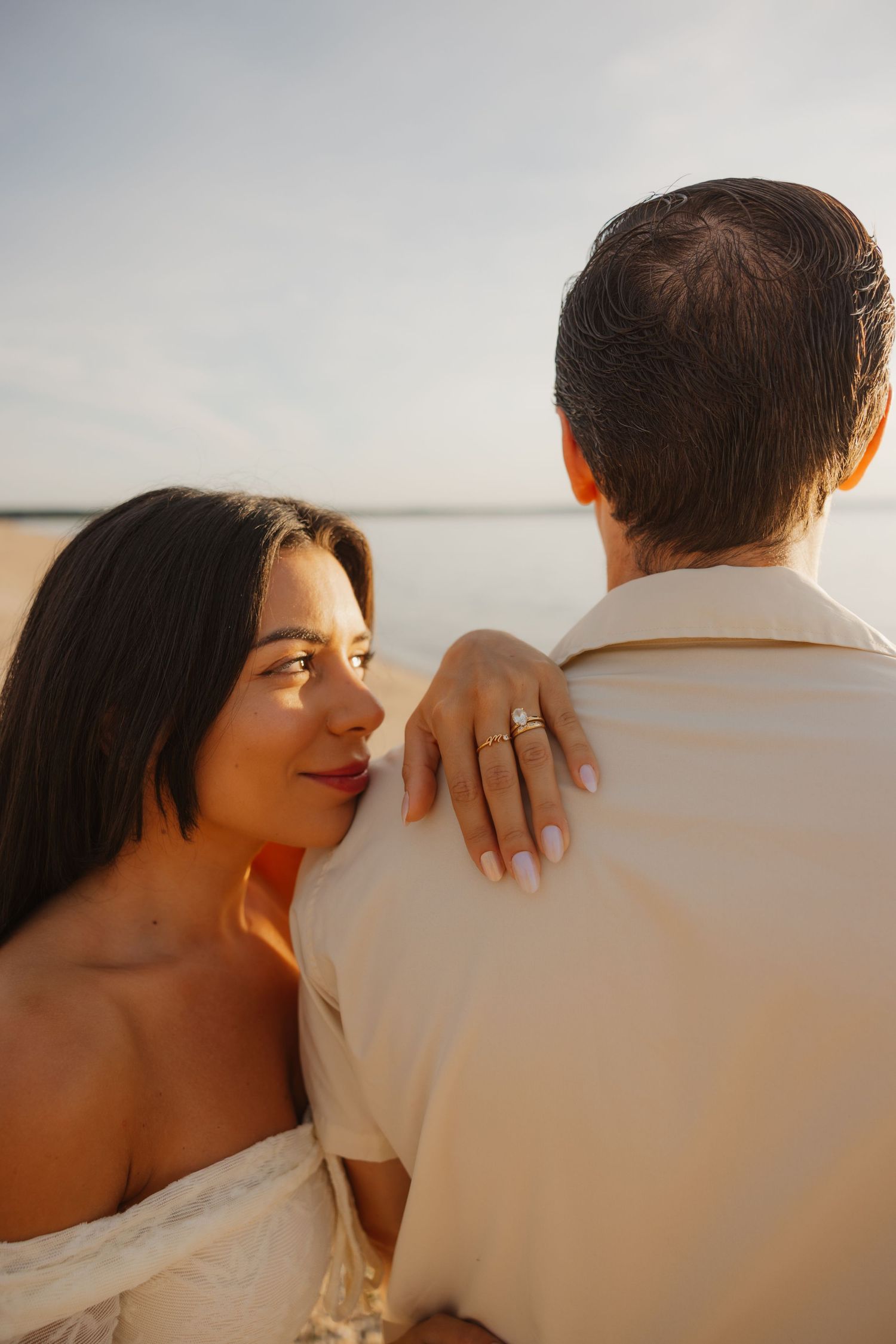 A woman with dark hair rests her hand on a man's shoulder, showing a gold ring, near a serene waterfront at sunset.