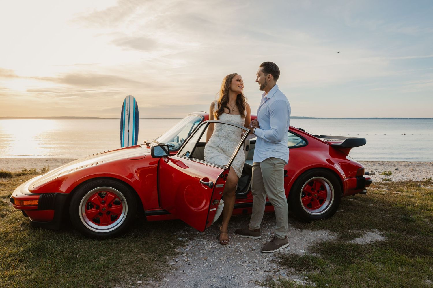 Couple posing beside a red Porsche 911 convertible with a surfboard at sunset near the ocean.