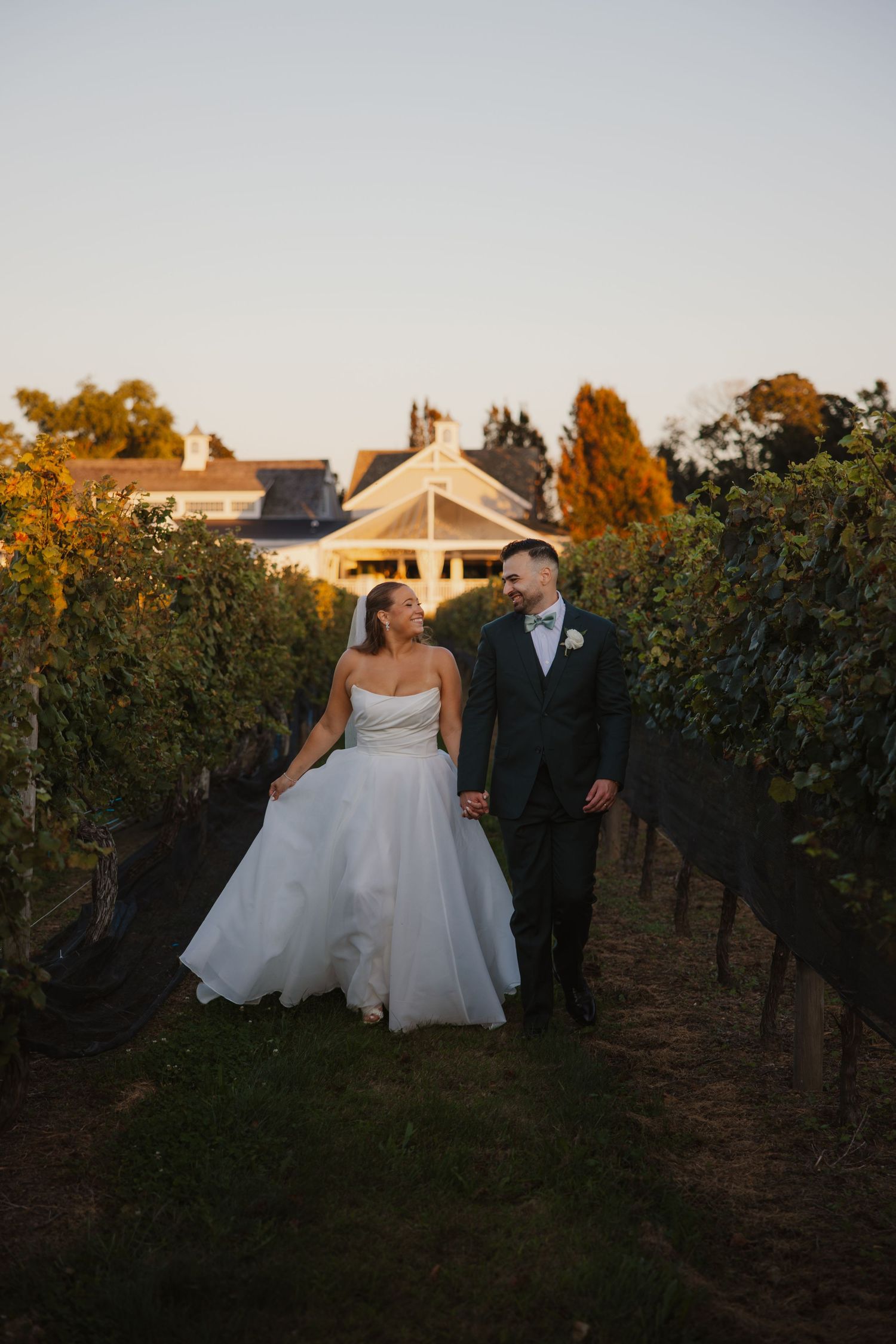 Bride and groom walk hand in hand through a vineyard at golden hour, with a white venue building in the background.