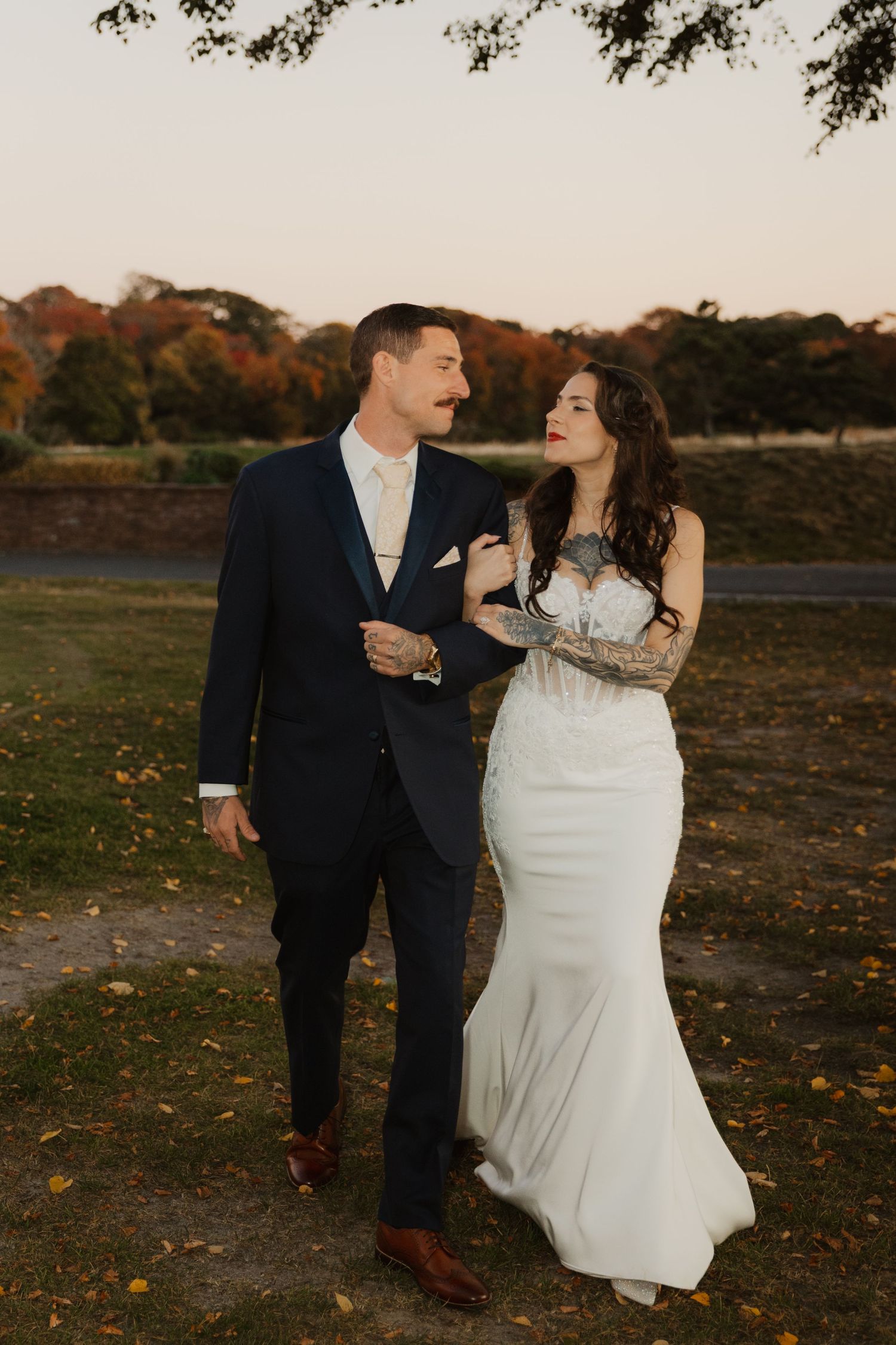 Bride in a fitted white lace gown walks arm in arm with groom in a navy suit across a grassy field with autumn trees at sunset.