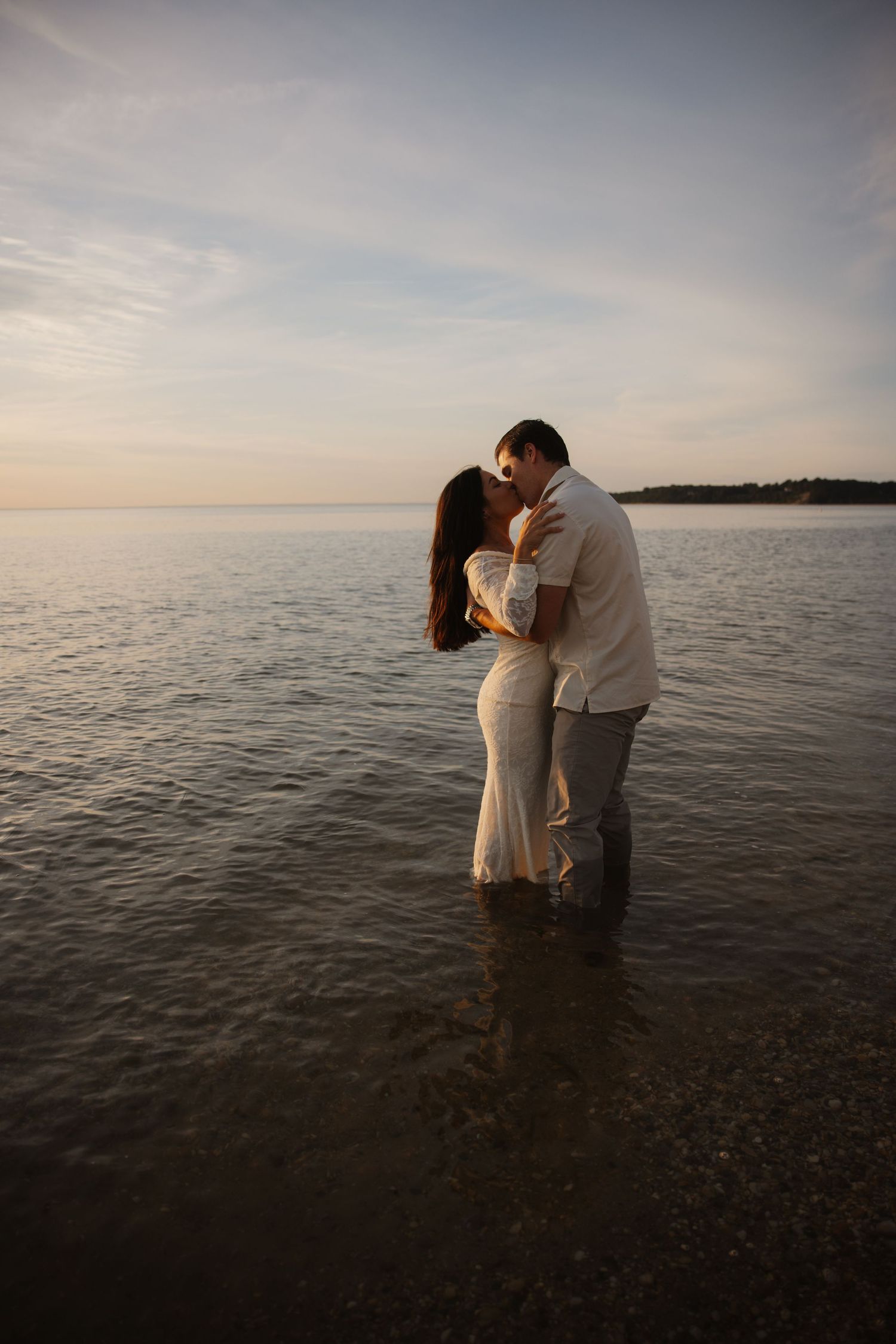 Couple kissing romantically while standing in shallow ocean water at sunset, woman in white dress, man in white shirt.