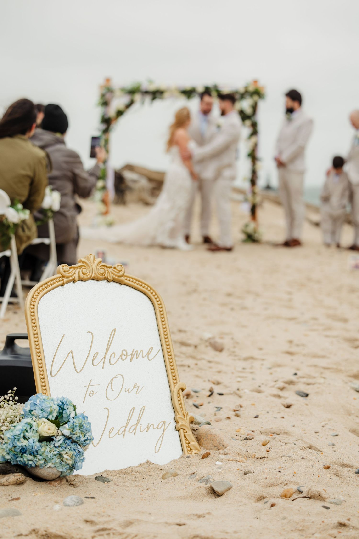 Welcome to Our Wedding sign in gold mirror frame at a beach ceremony with couple in background.