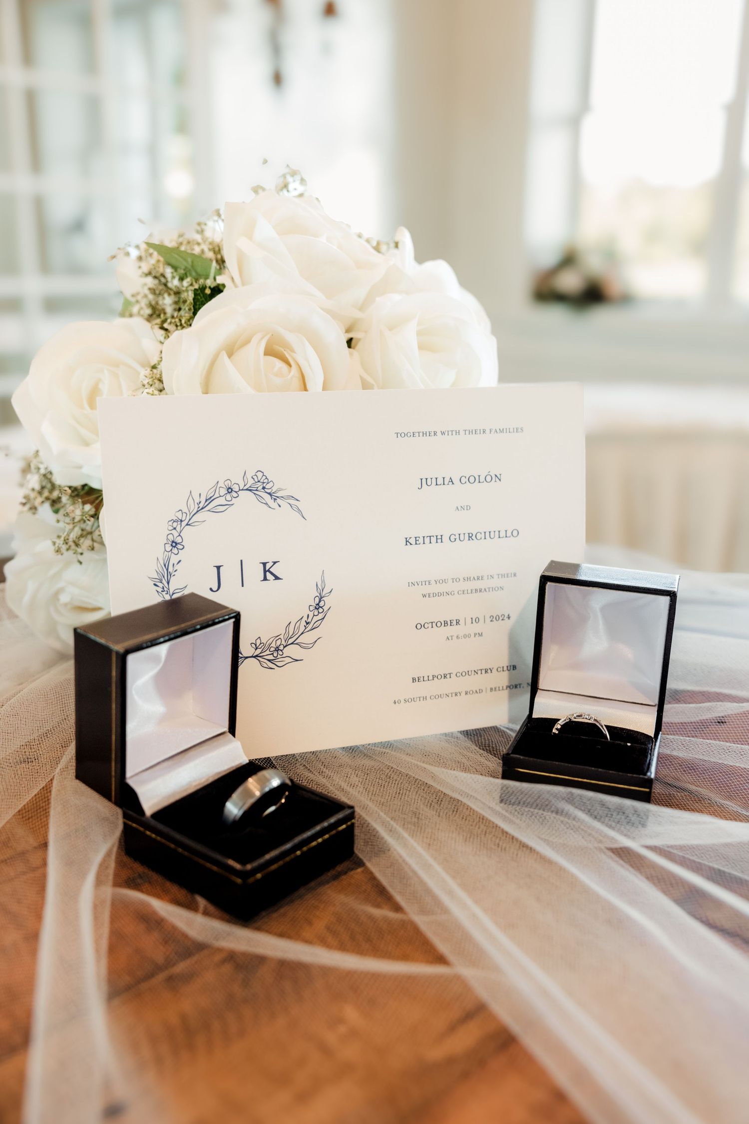 Elegant wedding flat lay with monogram invitation, white roses, and two ring boxes on a wooden table.
