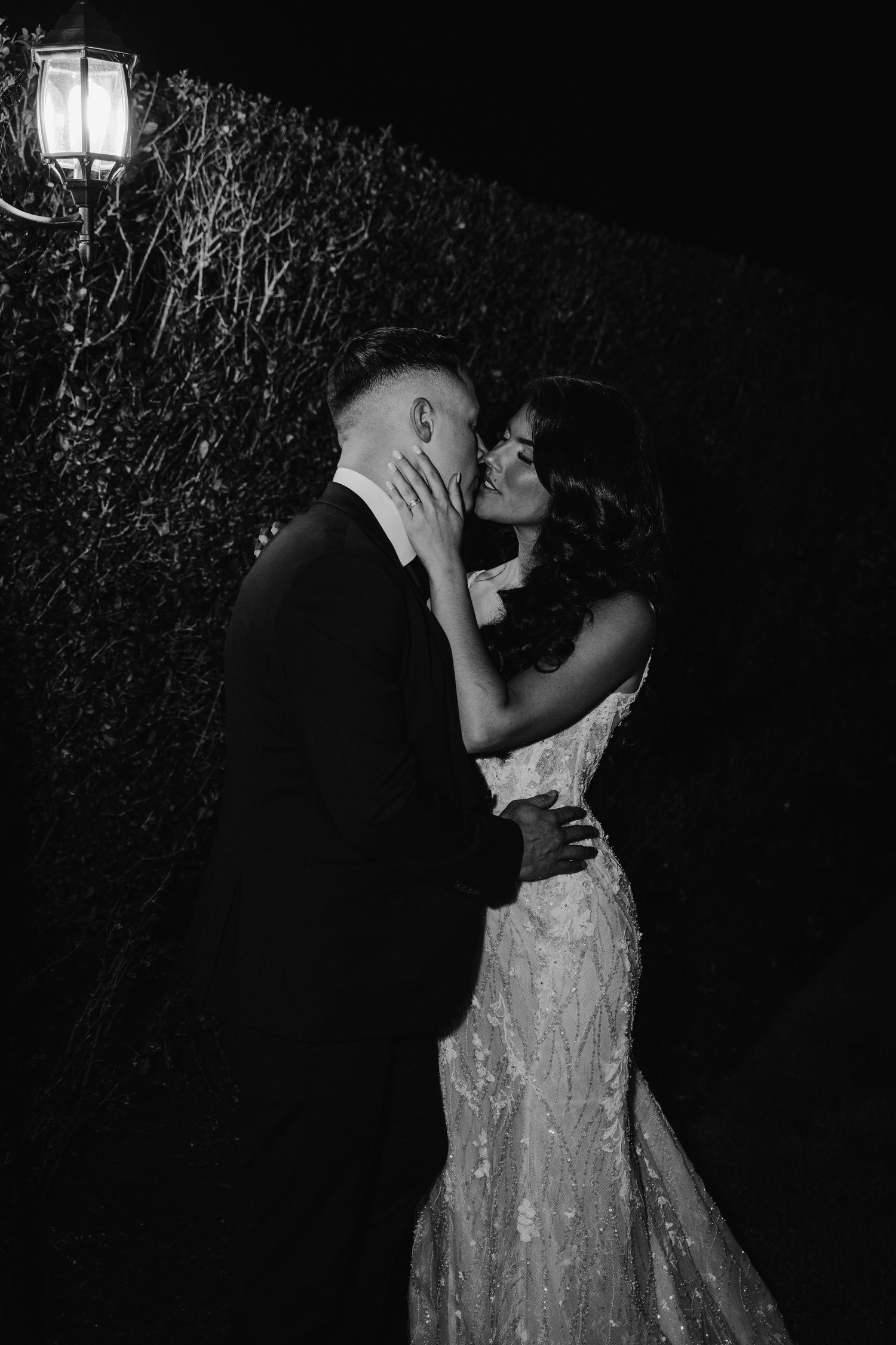 Black and white romantic photo of bride and groom sharing a kiss against an ivy-covered wall at night.