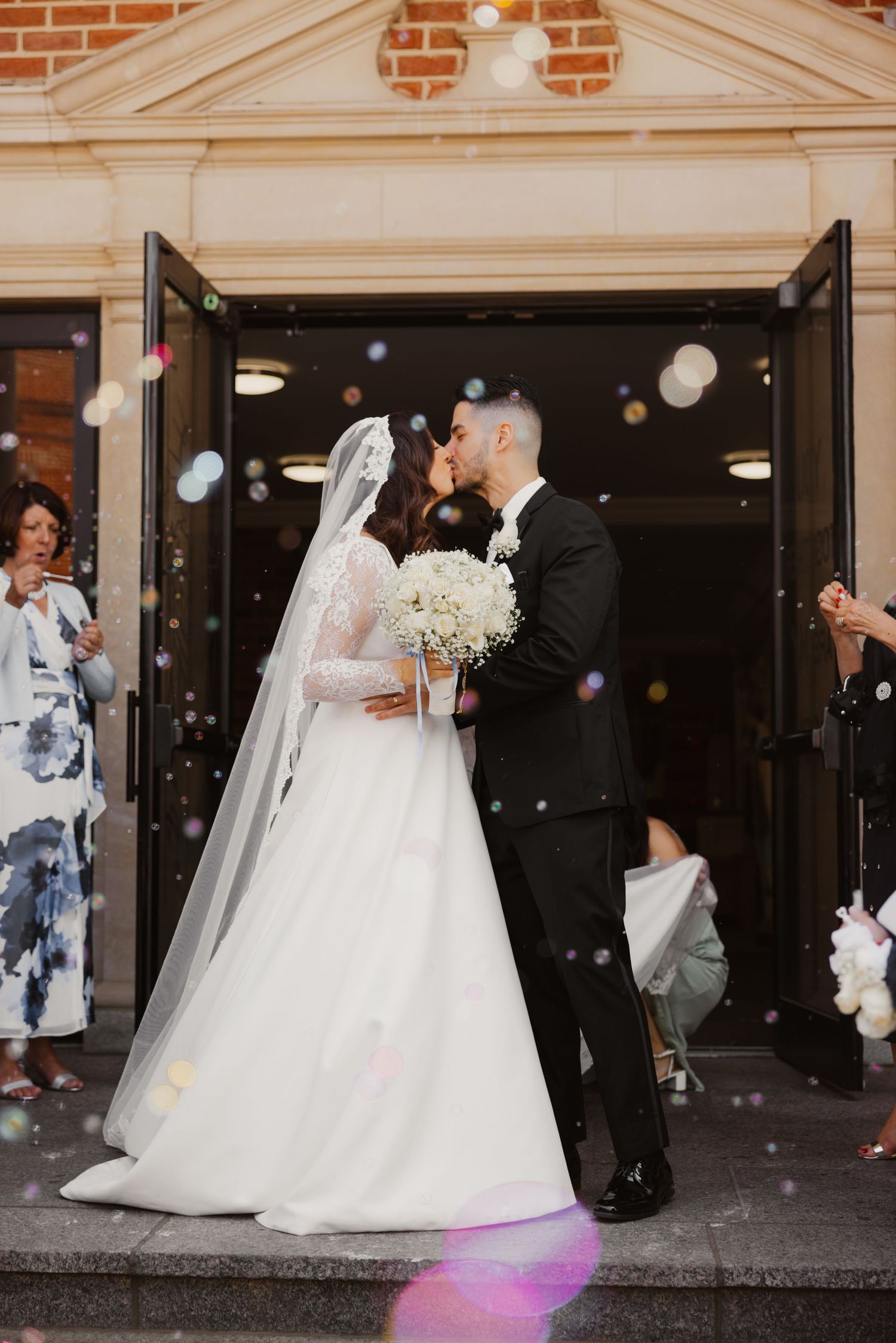 Newlyweds share a romantic kiss outside church doors surrounded by floating bubbles and guests.