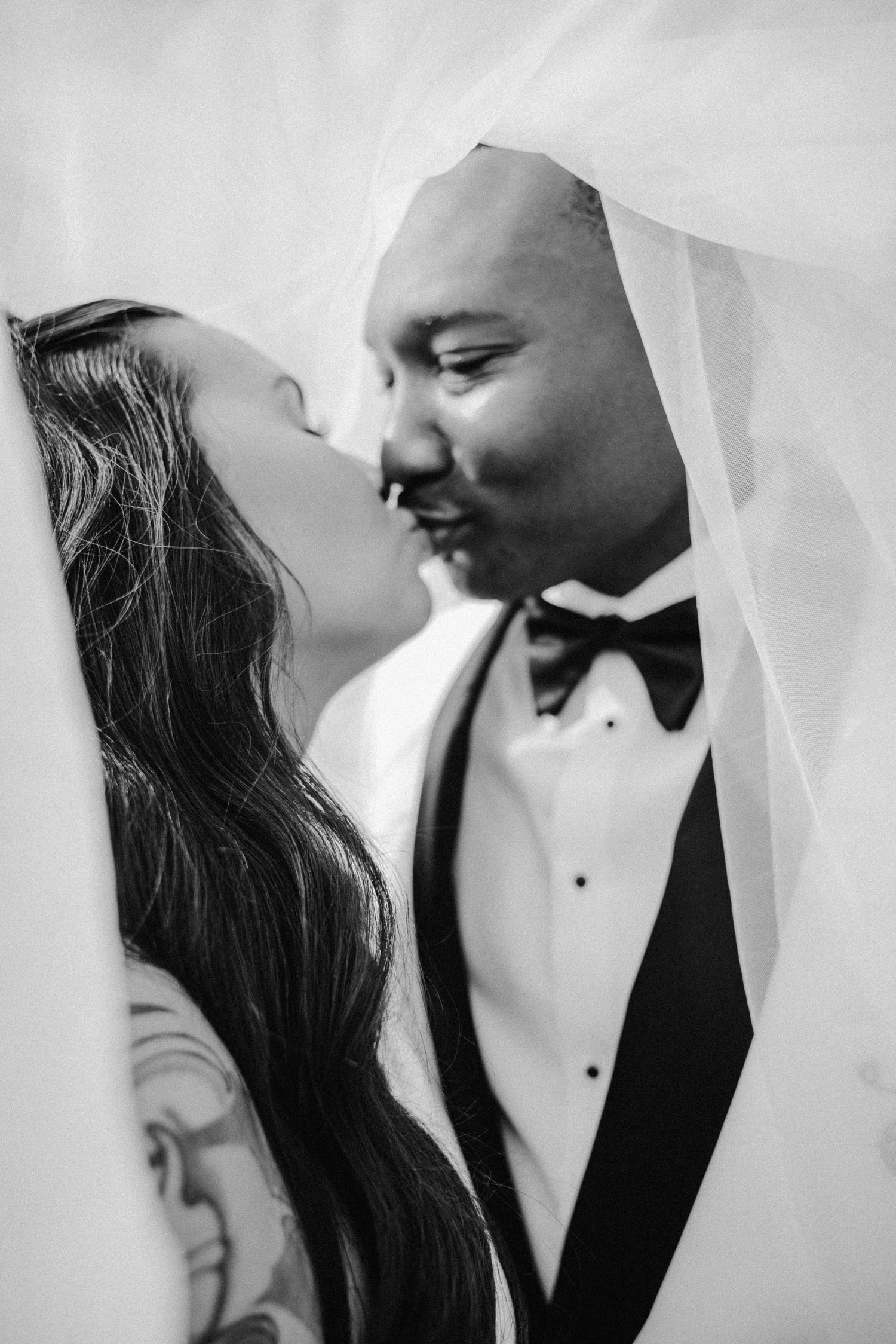 Black and white intimate portrait of bride and groom nearly kissing beneath a flowing wedding veil.