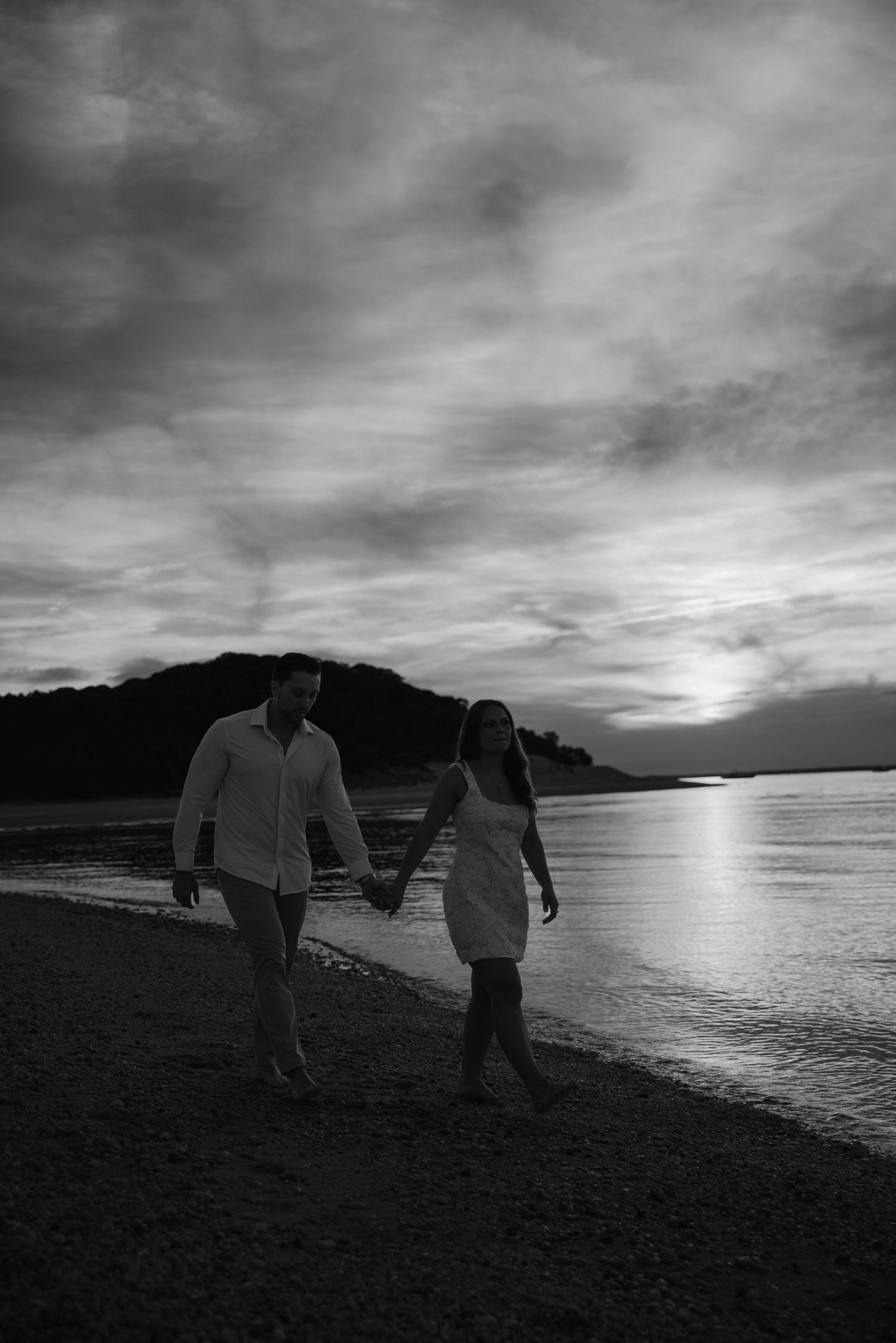 Black and white photo of a couple holding hands walking along a beach at sunset with dramatic cloudy sky.
