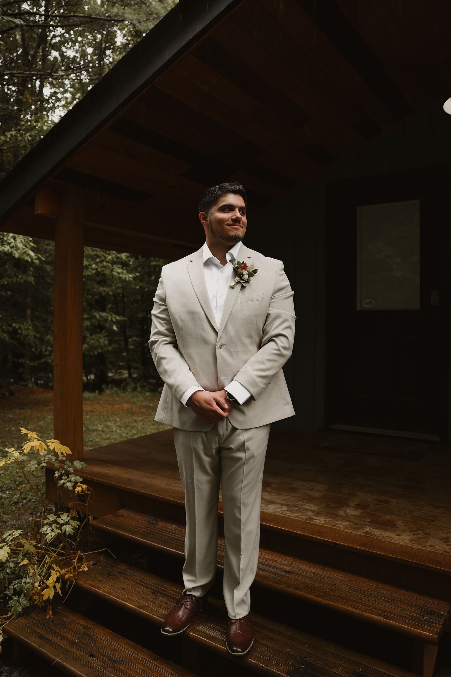 Dapper groom in beige suit with floral boutonniere stands on wooden cabin porch steps.