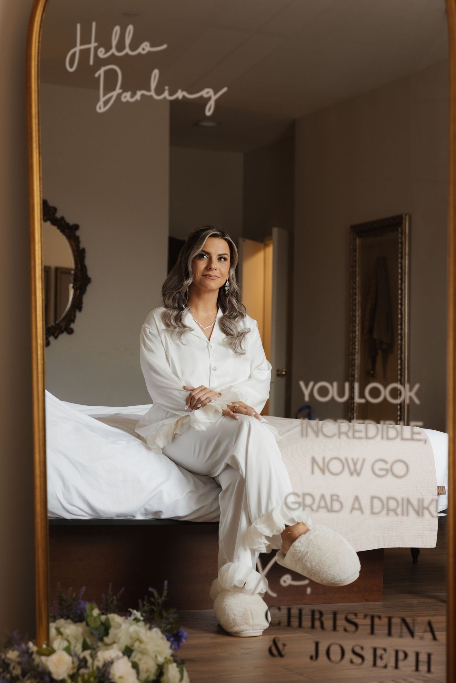 Bride sits on bed in white satin pajamas with fluffy slippers in elegant hotel room before wedding.