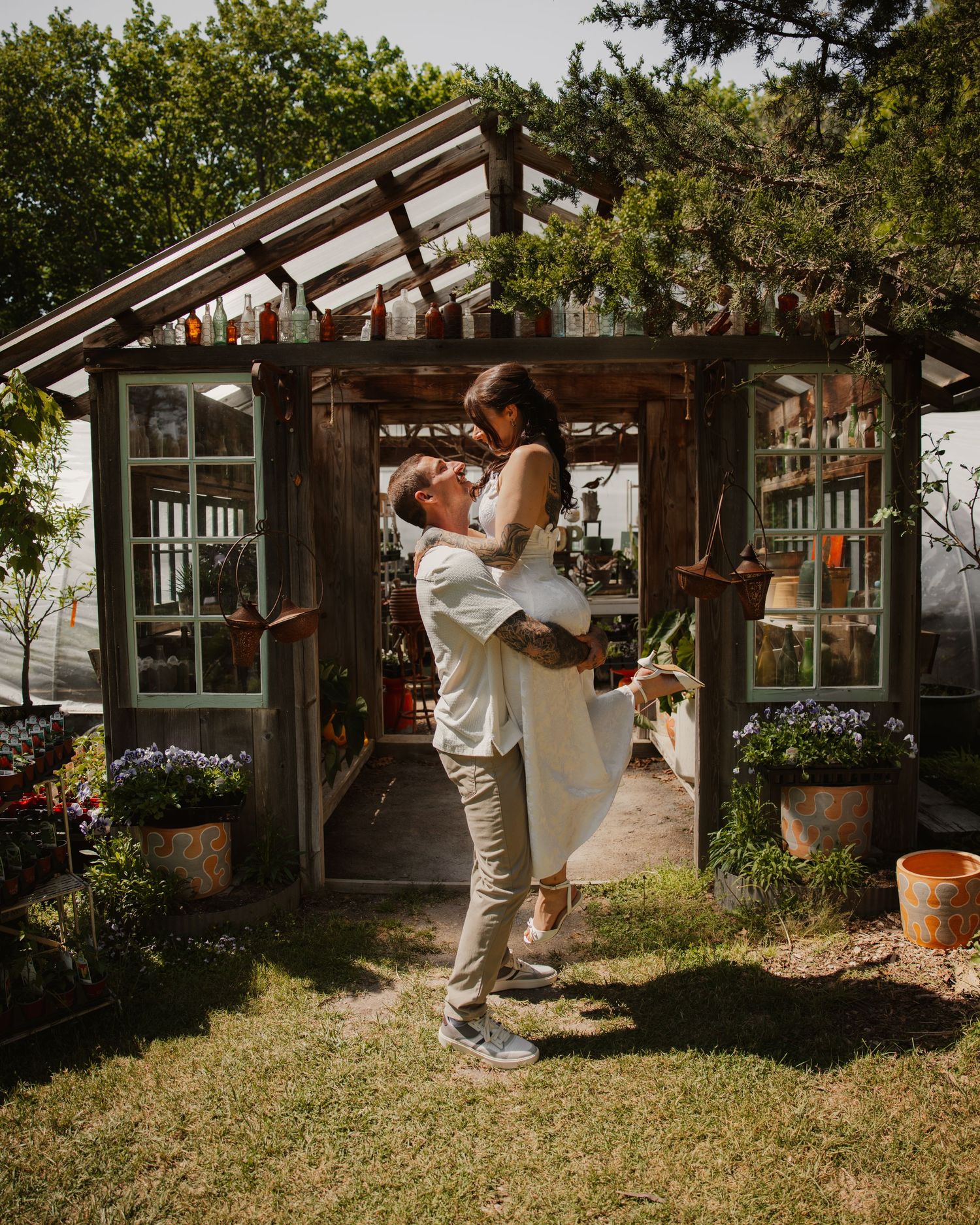 Groom lifts bride in front of a rustic wooden greenhouse surrounded by potted plants.