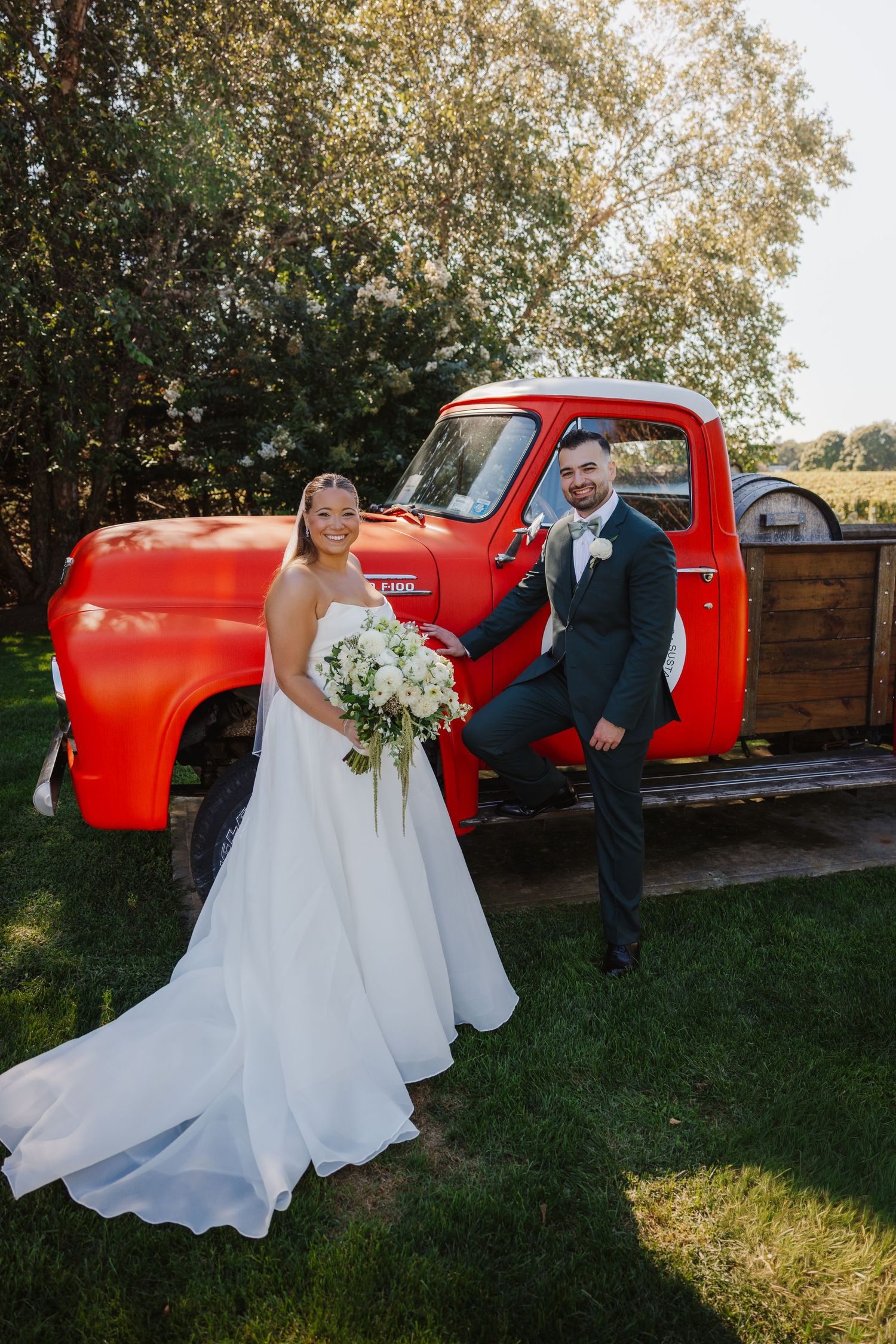 Bride and groom pose smiling in front of a classic red truck on a sunny day, bride holding a white bouquet.