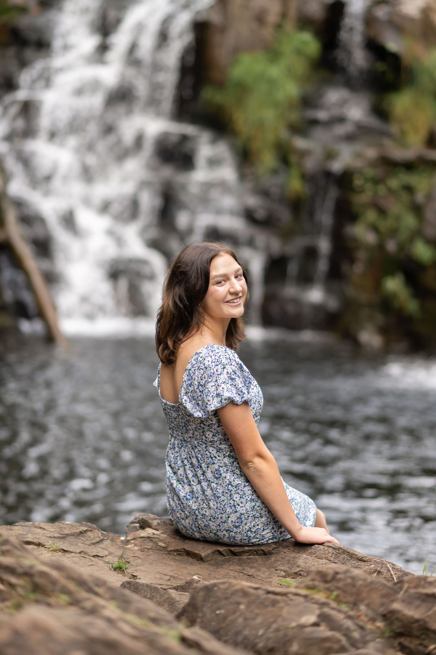 High school senior girl sitting on rocks beside a waterfall during a scenic outdoor senior photography session in Washington.